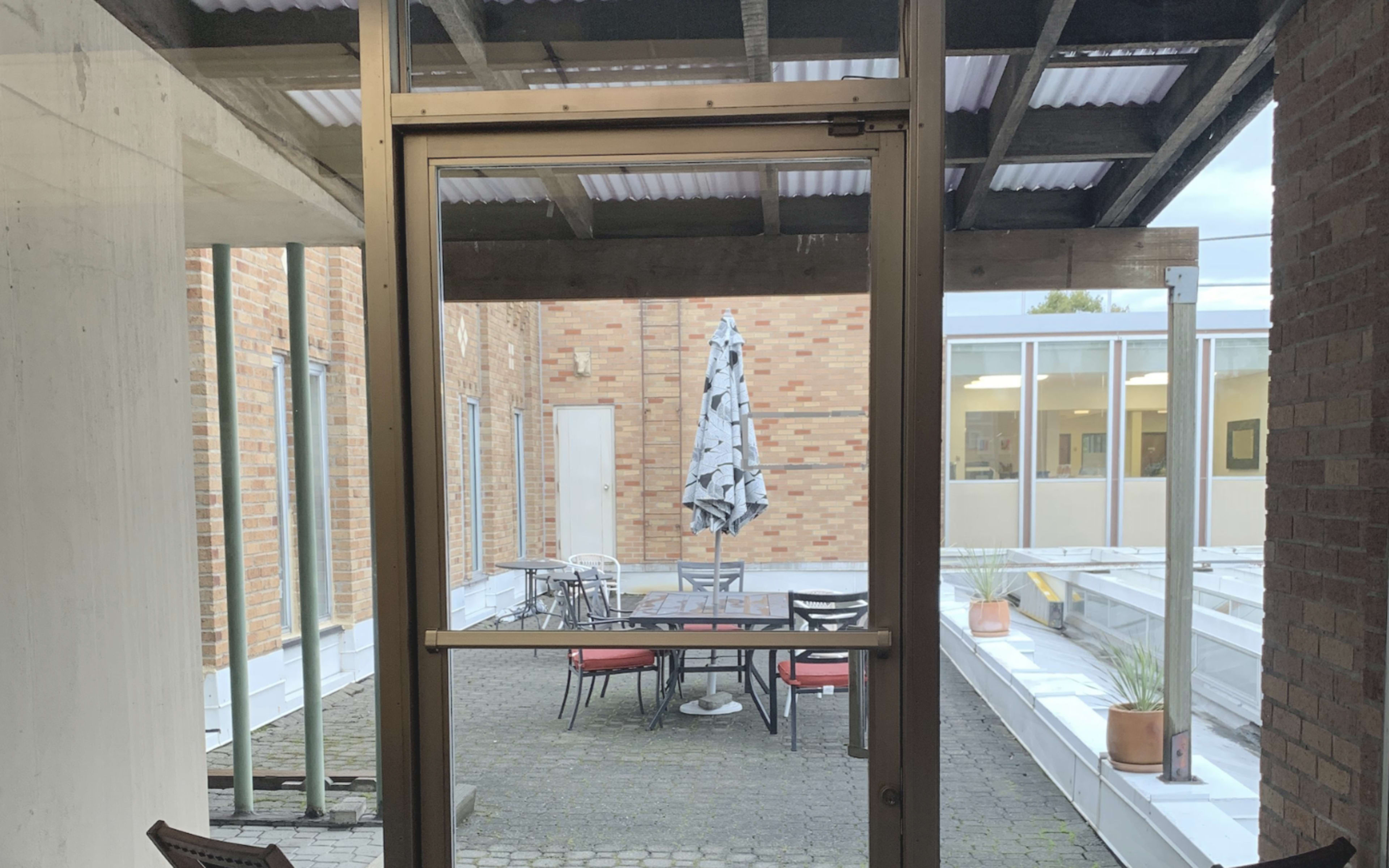 The image shows a view through a glass door into a courtyard featuring a table with chairs and a large umbrella, surrounded by brick and paved surfaces.