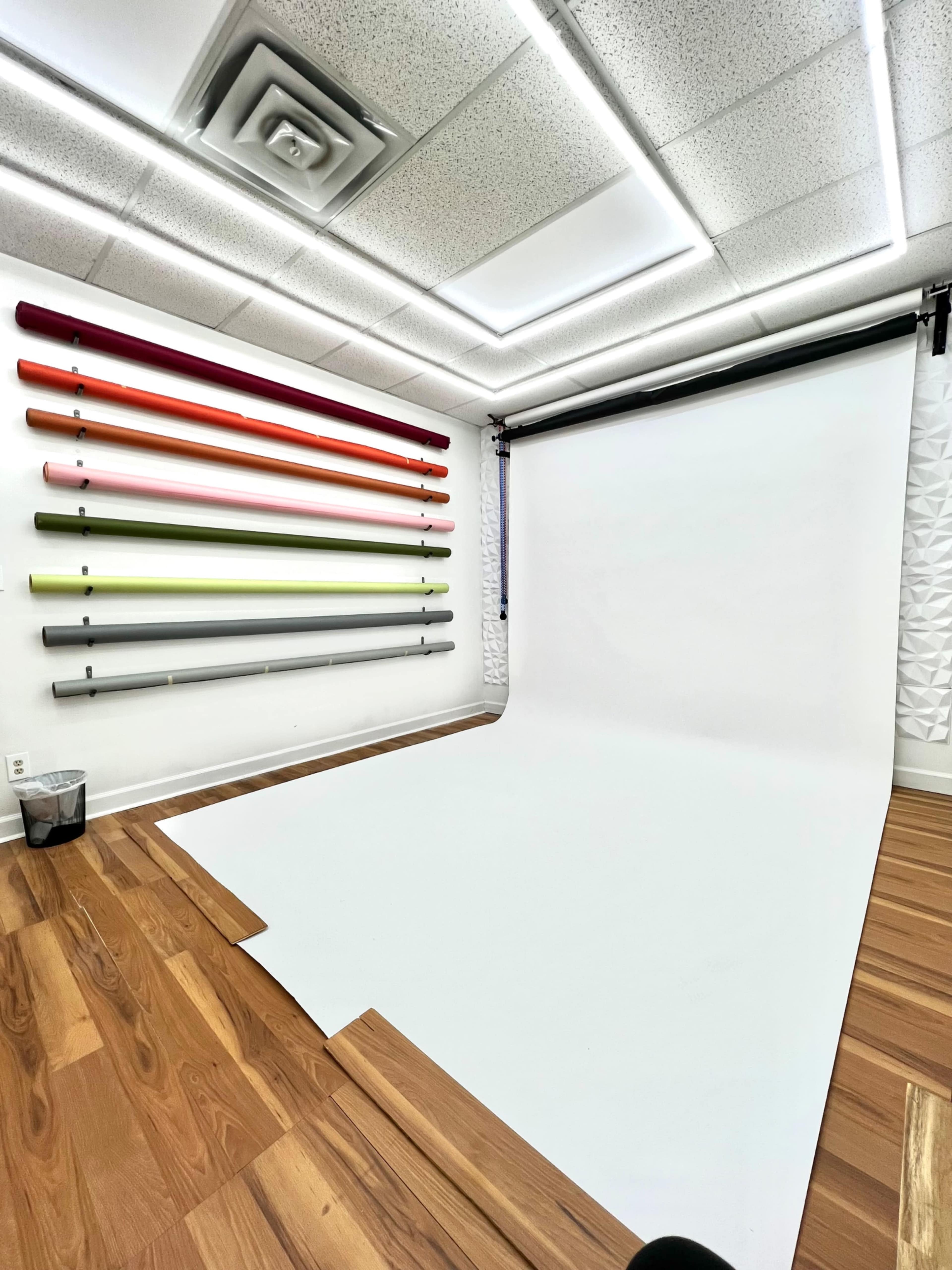 The image shows a modern studio space with a white backdrop, wooden flooring, and a wall featuring colorful rolls of paper arranged horizontally.