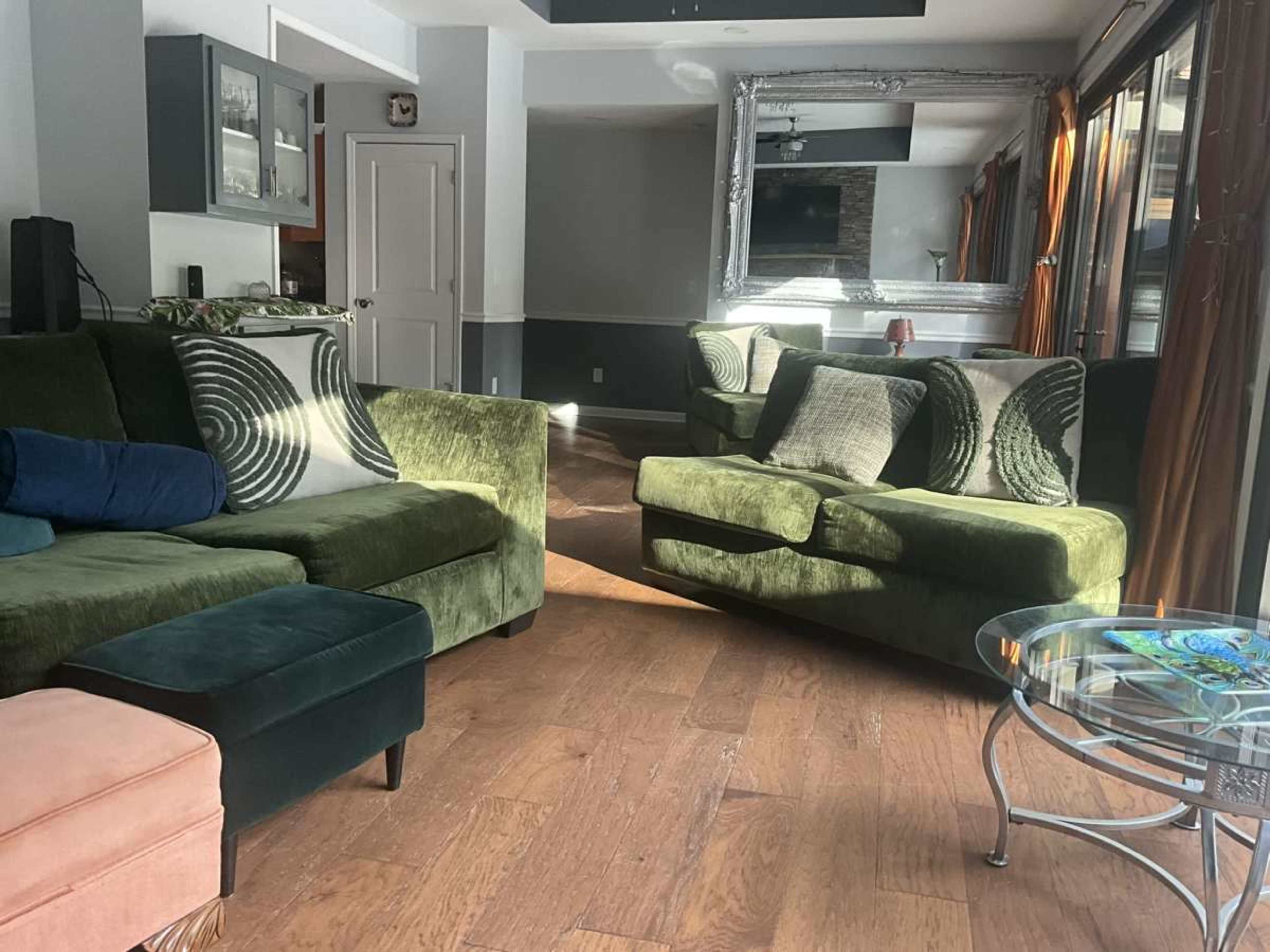 The image shows a cozy living room with green sofas, a glass coffee table, and large windows letting in natural light.