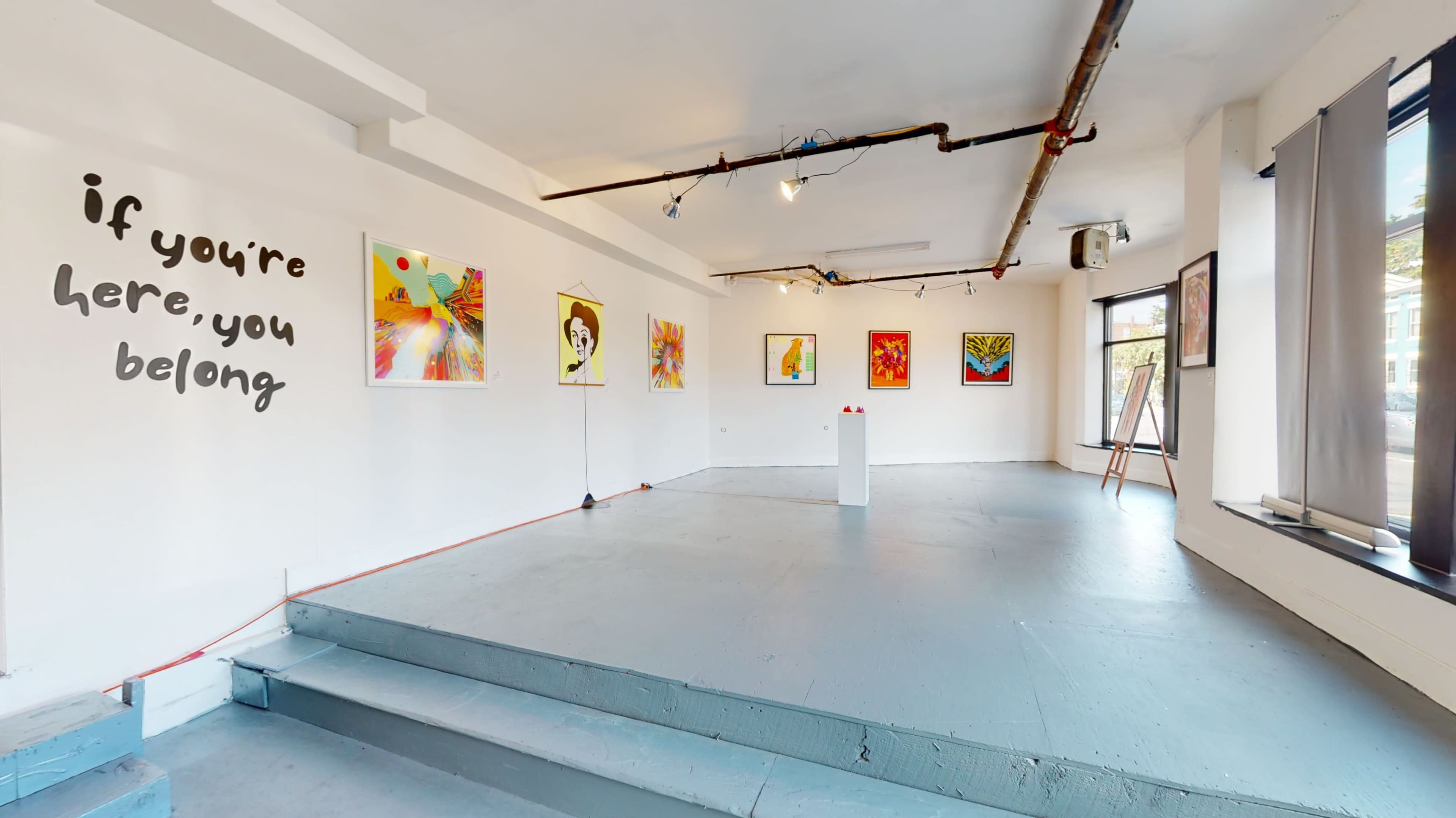 A spacious art gallery with colorful paintings displayed on the white walls and a minimalist pedestal in the center.