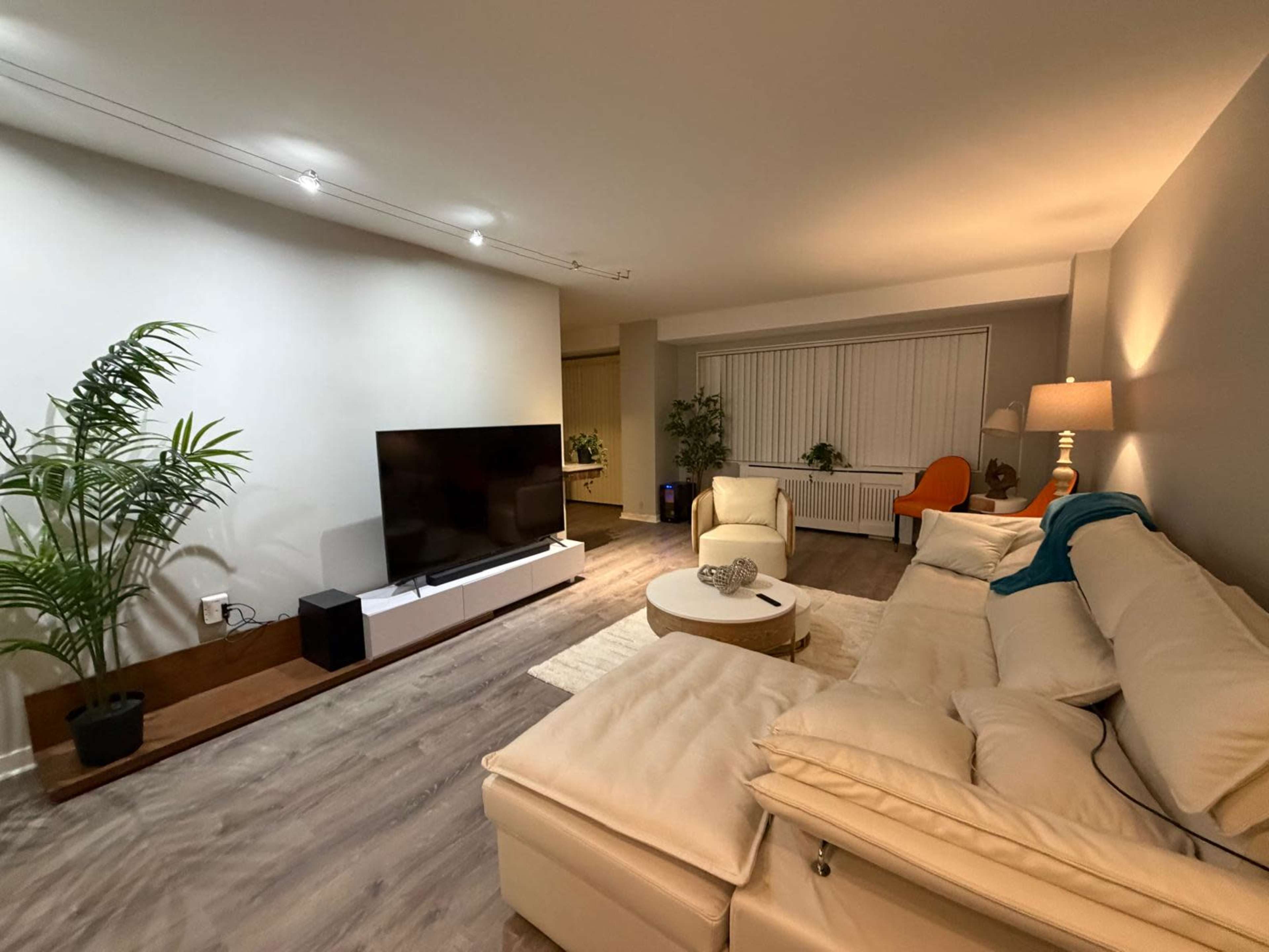The image shows a modern living room with a sectional sofa, a large television on a stand, a potted plant, and a cozy ambiance created by soft lighting.