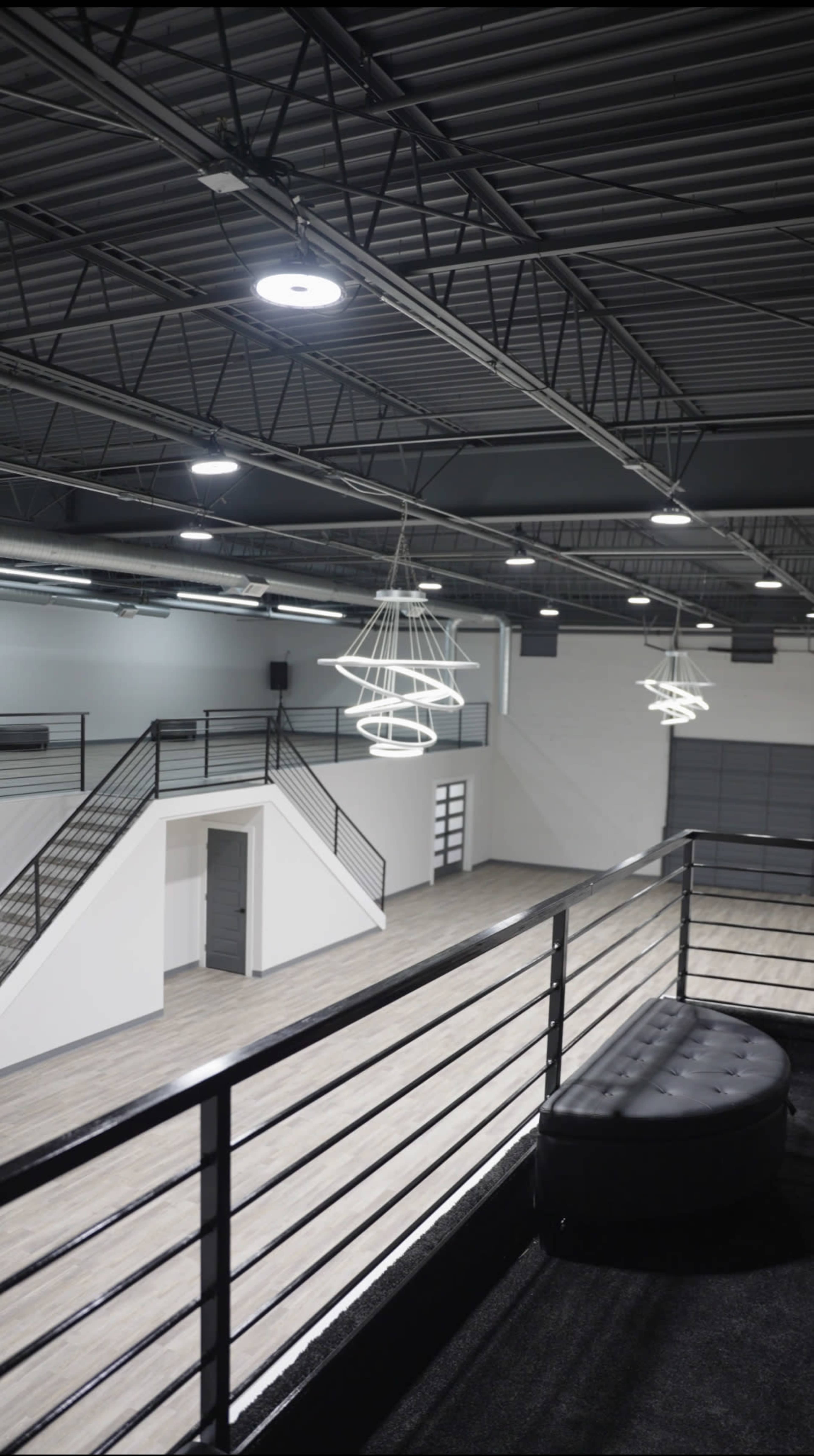 The image shows a spacious, modern interior with high ceilings, featuring sleek lighting fixtures, a staircase, and a large open area with wooden flooring.