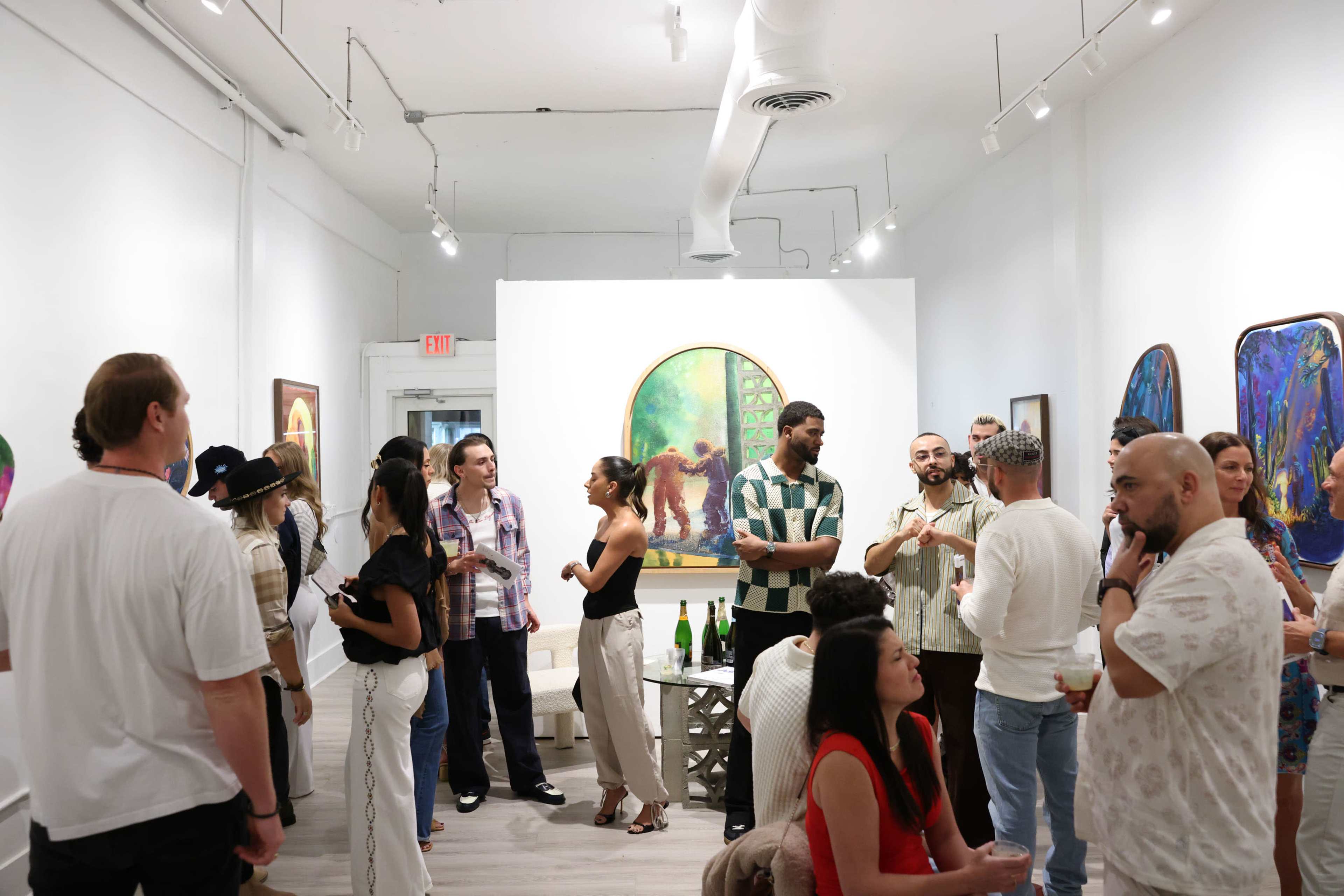 A group of people is conversing and mingling in an art gallery featuring colorful paintings on the walls.