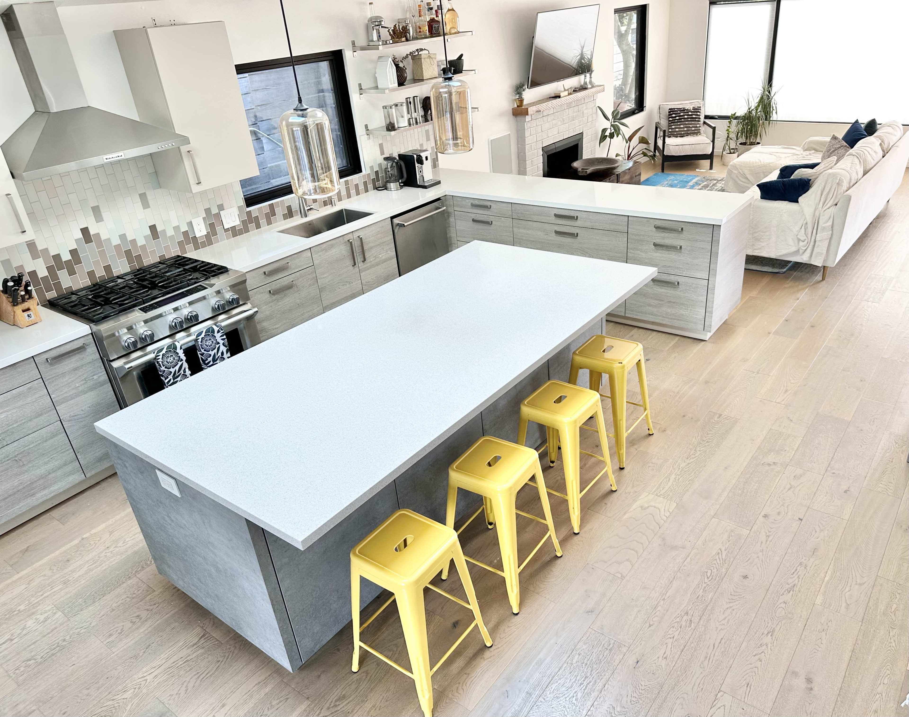 A modern kitchen features a large gray island with four yellow stools and an open living area in the background.