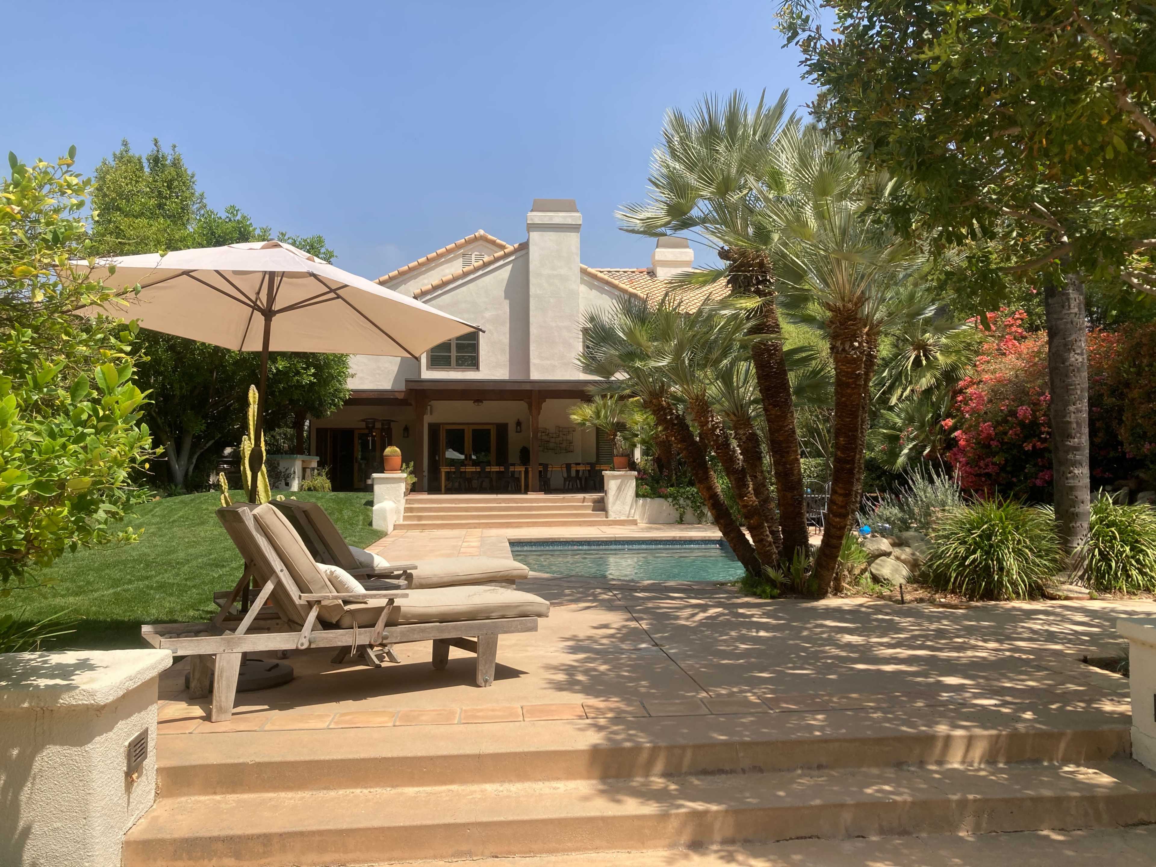 A shaded patio area with lounge chairs overlooks a swimming pool adjacent to a house surrounded by palm trees and lush landscaping.