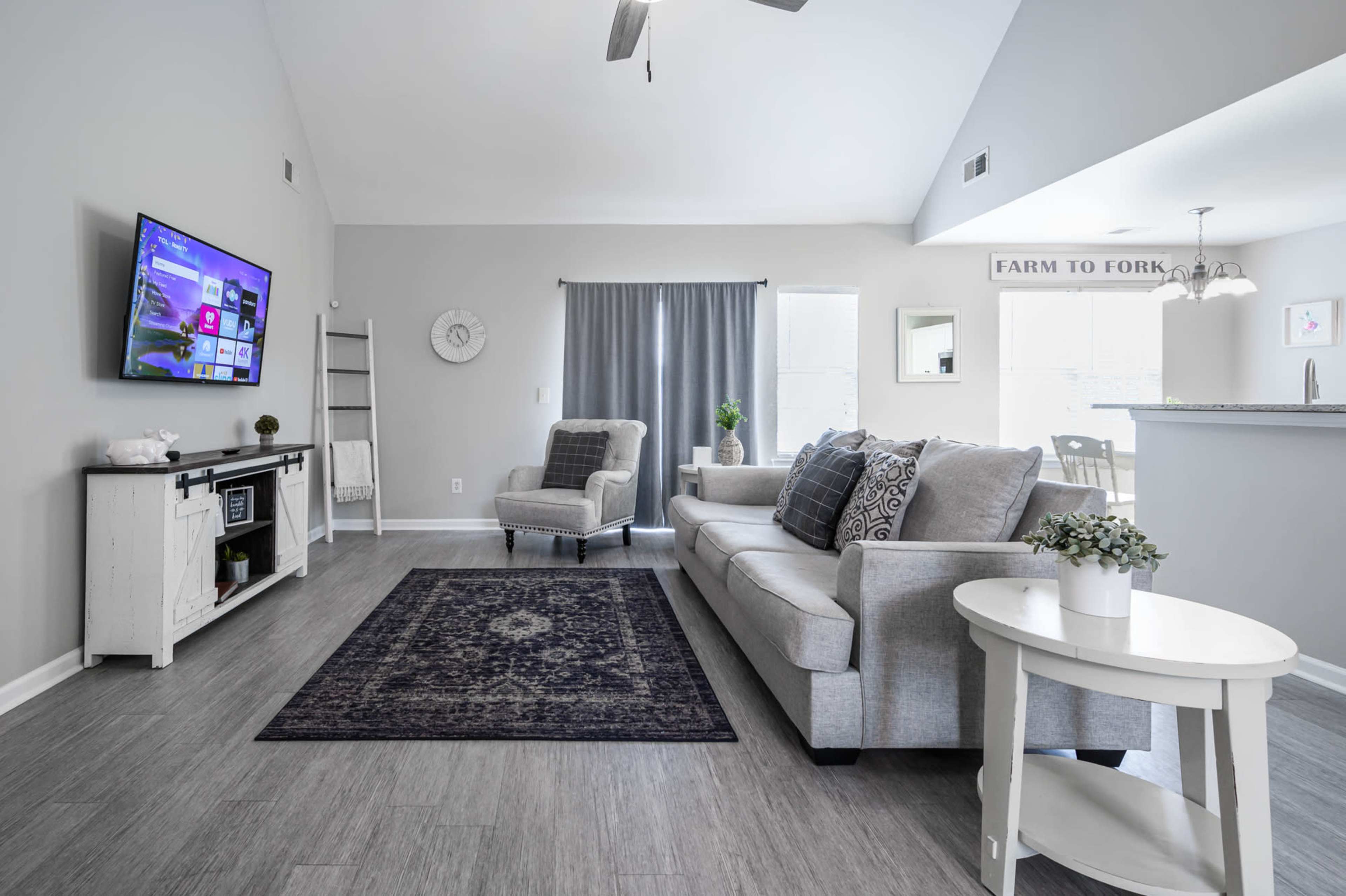 A modern living room features a gray sofa and a television mounted on the wall, with a dining area visible in the background.