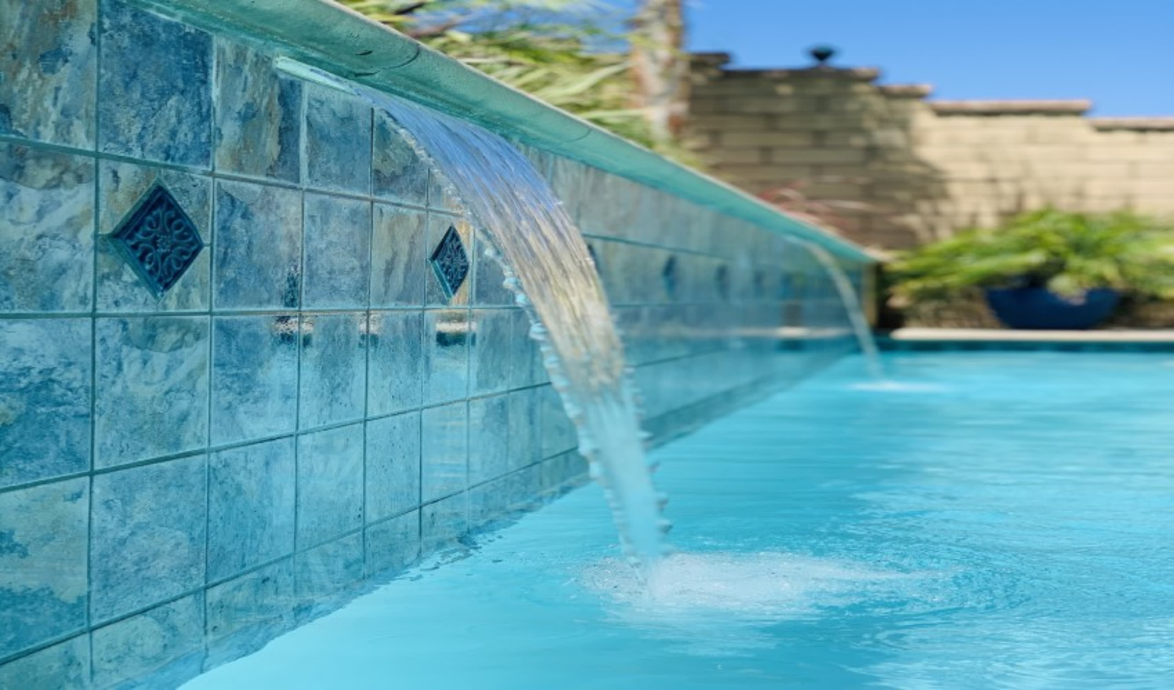 Water cascades from a tile wall into a swimming pool under a clear blue sky.