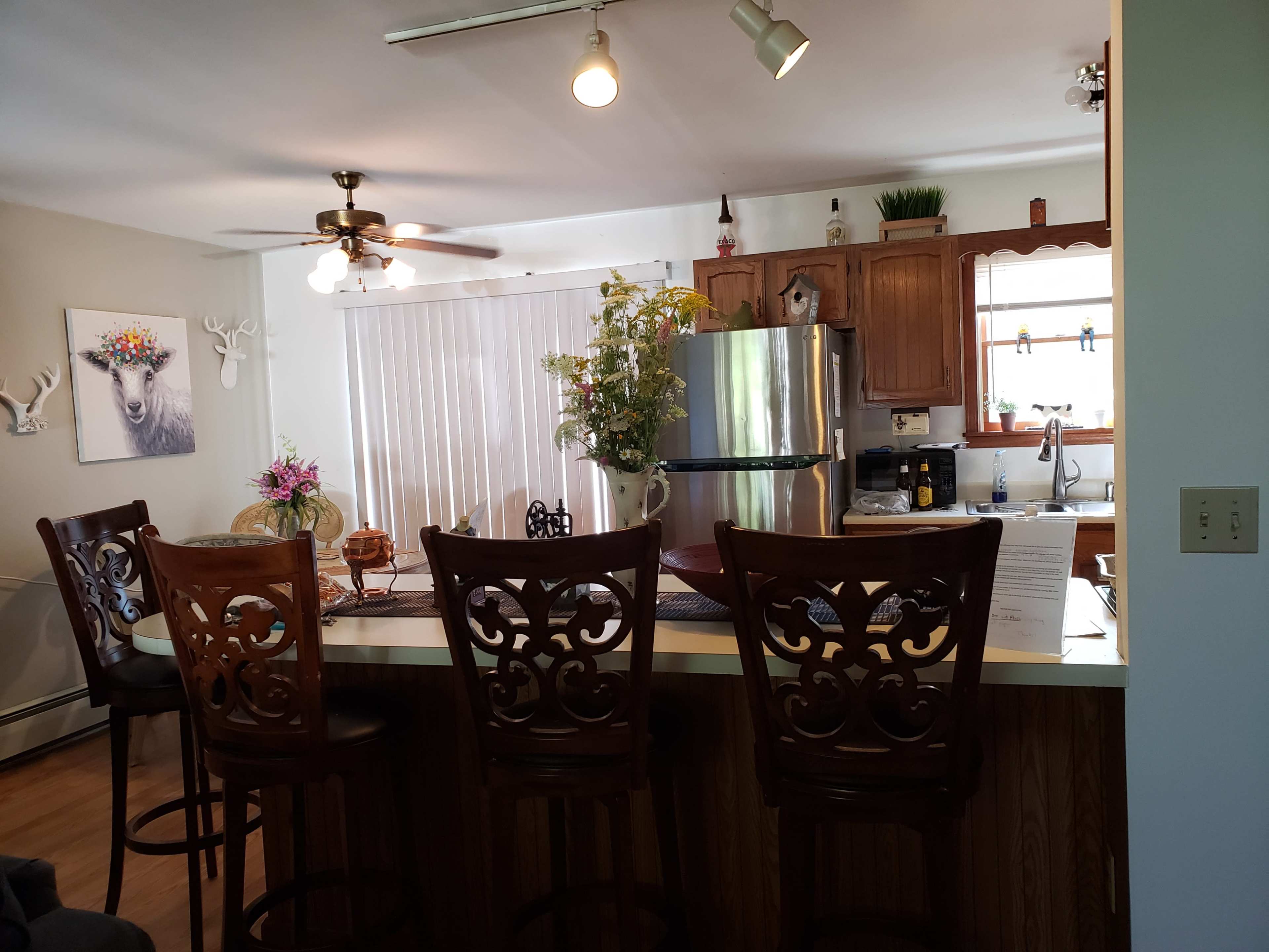 A kitchen features a bar with high stools, a stainless steel refrigerator, and a window overlooking a light-filled area with decorative plants and a deer-themed wall art.
