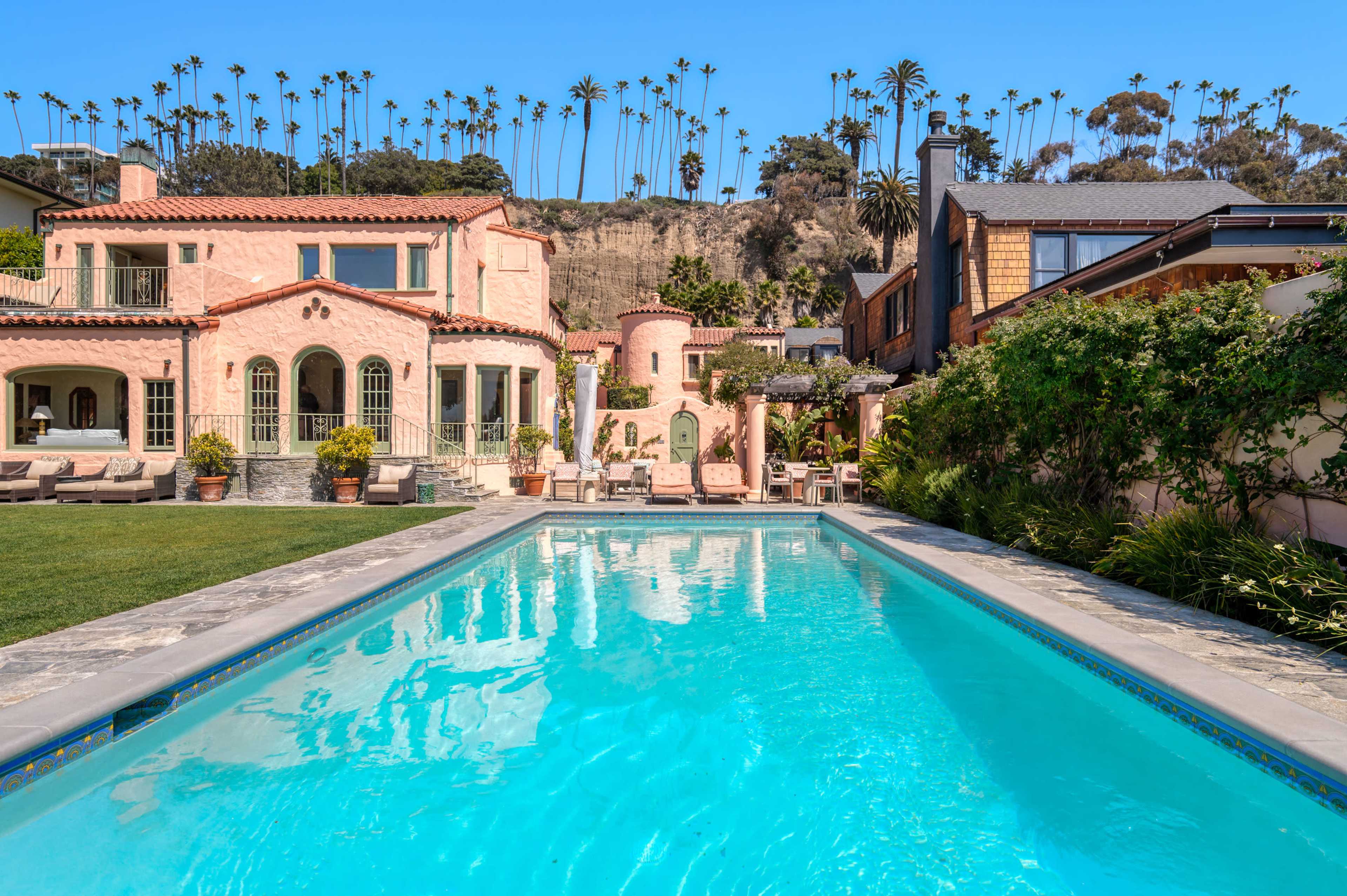 A modern home with a swimming pool in the foreground, surrounded by palm trees and terraced landscaping.