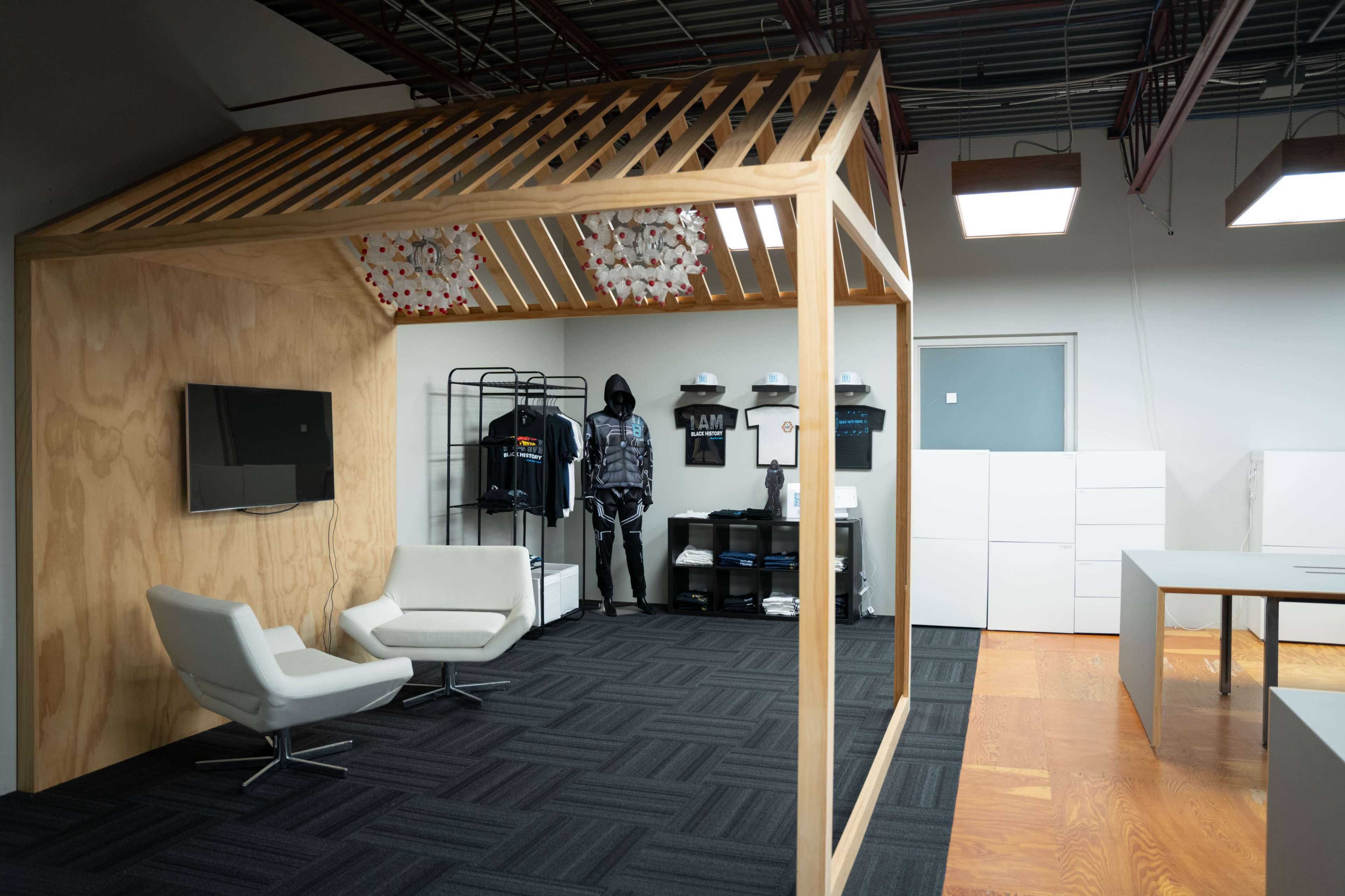 The image shows a modern office space featuring a wooden structure with a slatted roof, two white chairs, a television mounted on the wall, and a mannequin dressed in black attire standing near clothing displays.