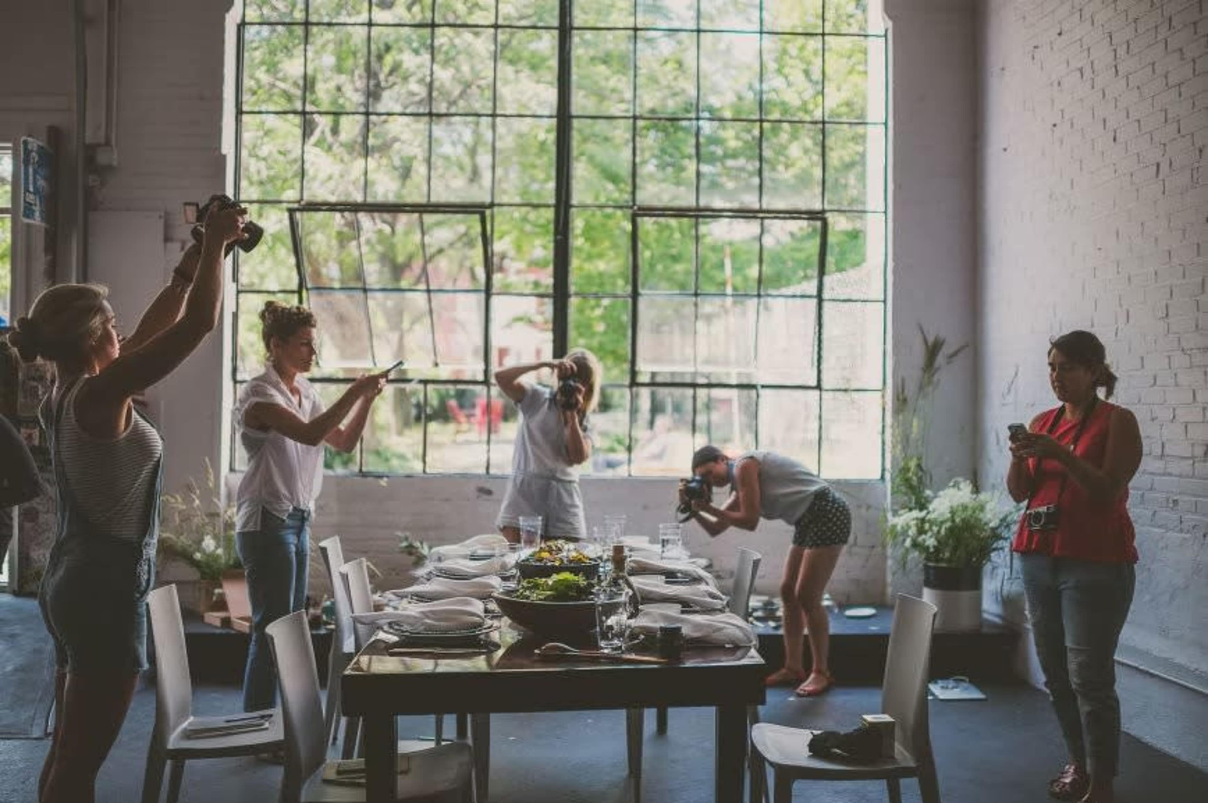 A group of six women are photographing a beautifully set dining table in a well-lit industrial space with large windows.