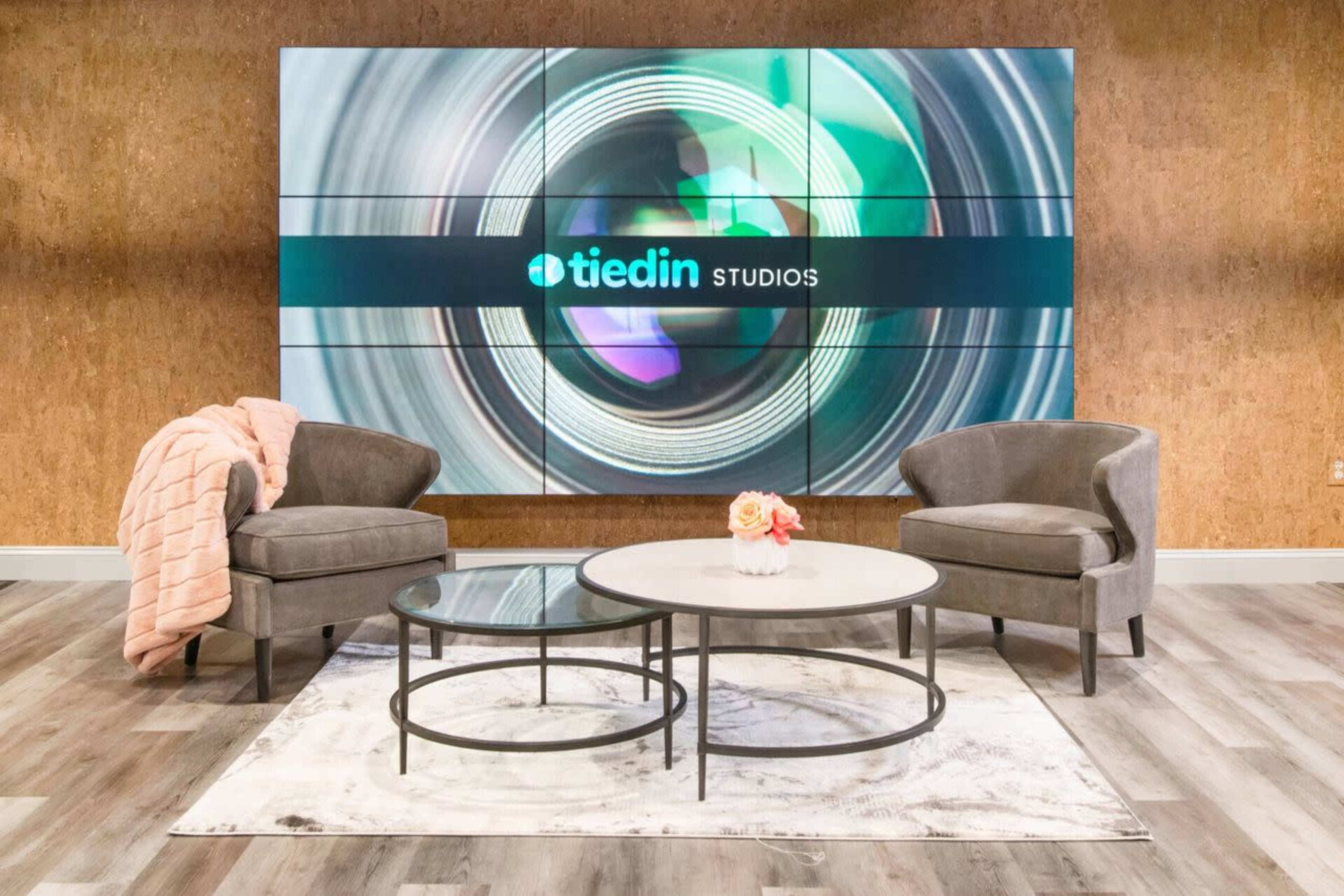 The image shows a modern studio setting featuring two gray armchairs, a round coffee table, and a large screen displaying the logo for "tiedin STUDIOS" against a textured wall.