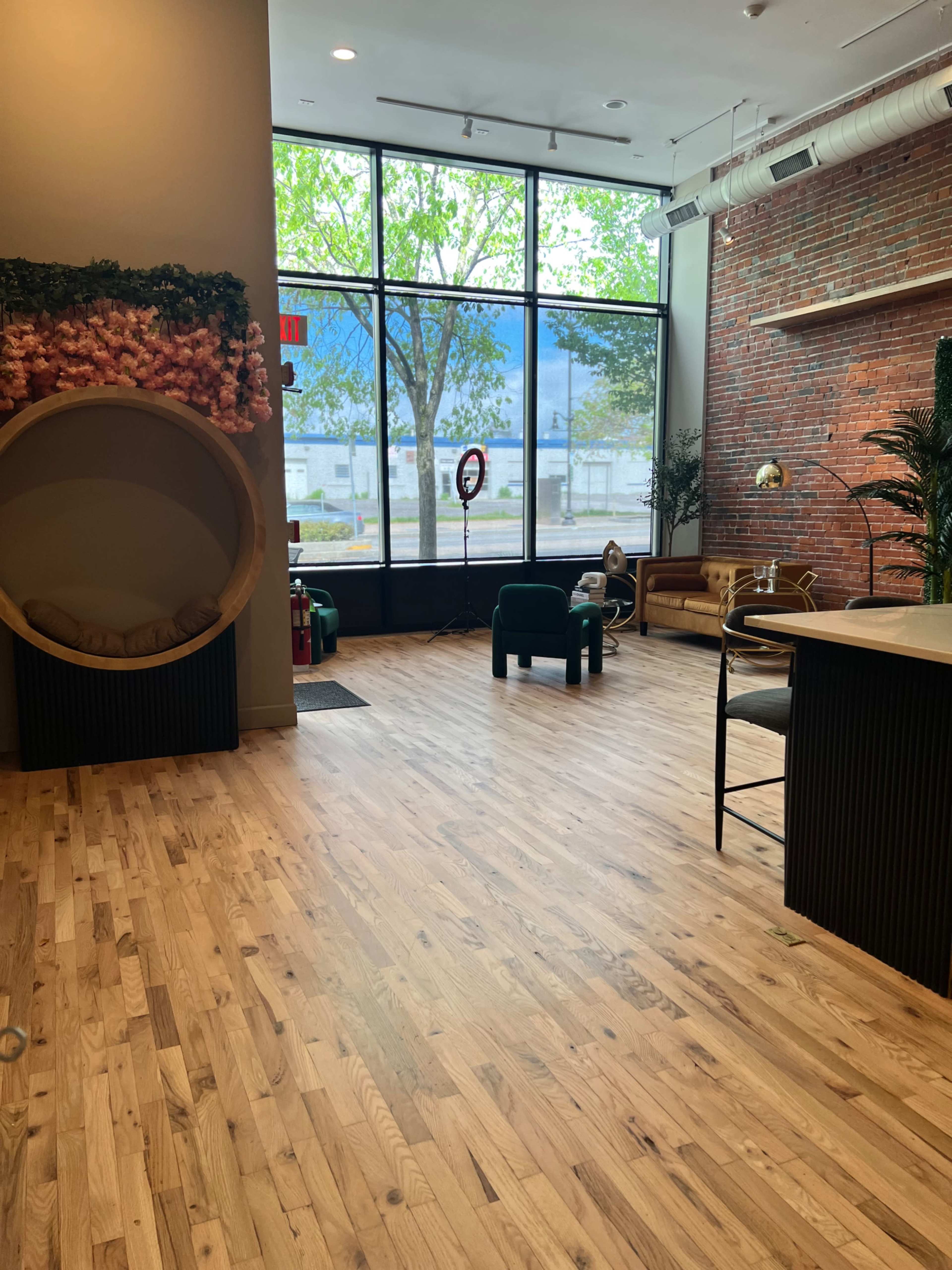 A spacious interior with wooden flooring, large windows letting in natural light, and a mix of furniture including a circular seating area, chairs, and decorative plants against a brick wall.