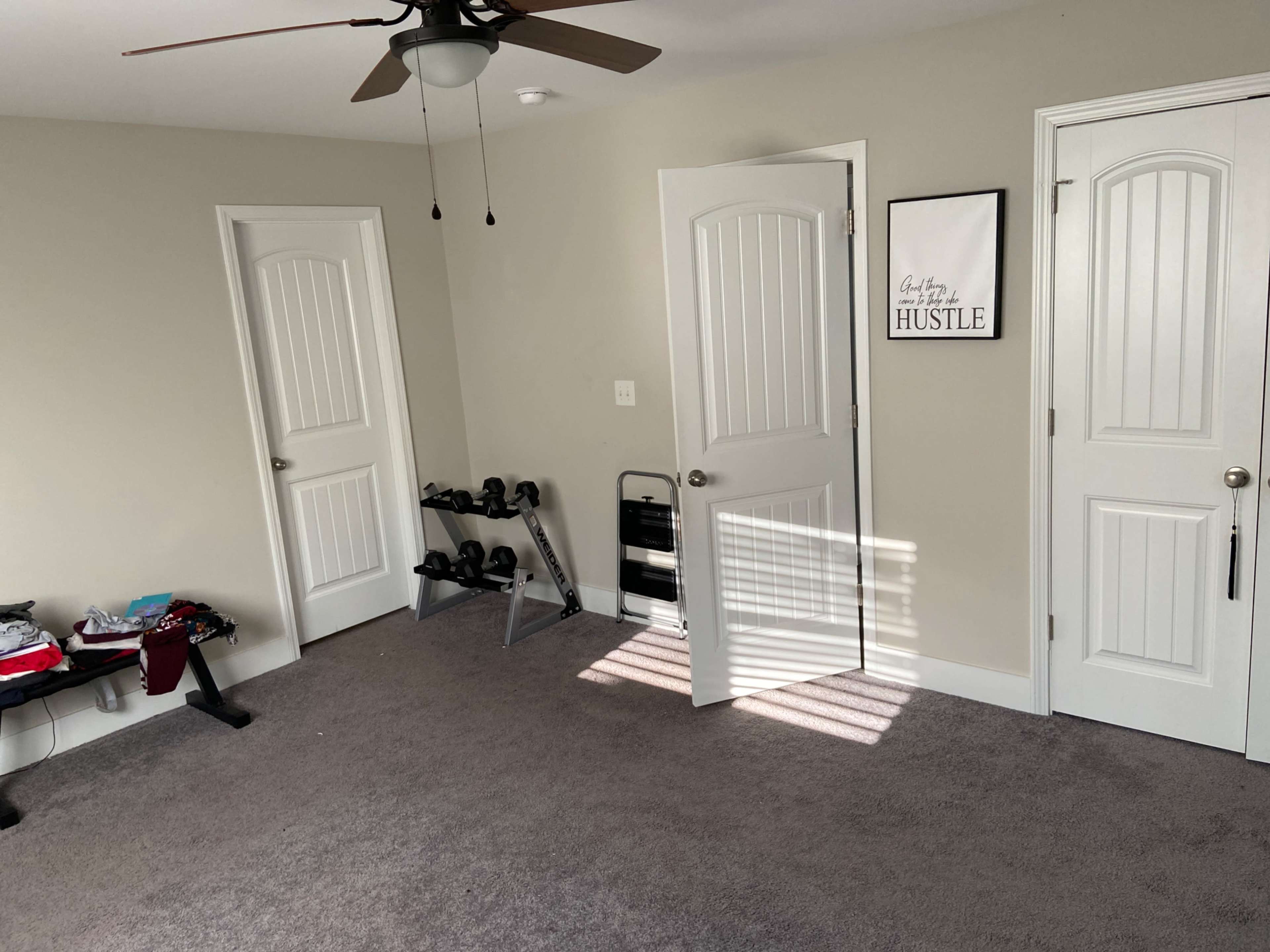A sparsely furnished room with two closed doors, a wall-mounted fan, and a small exercise area featuring dumbbells.