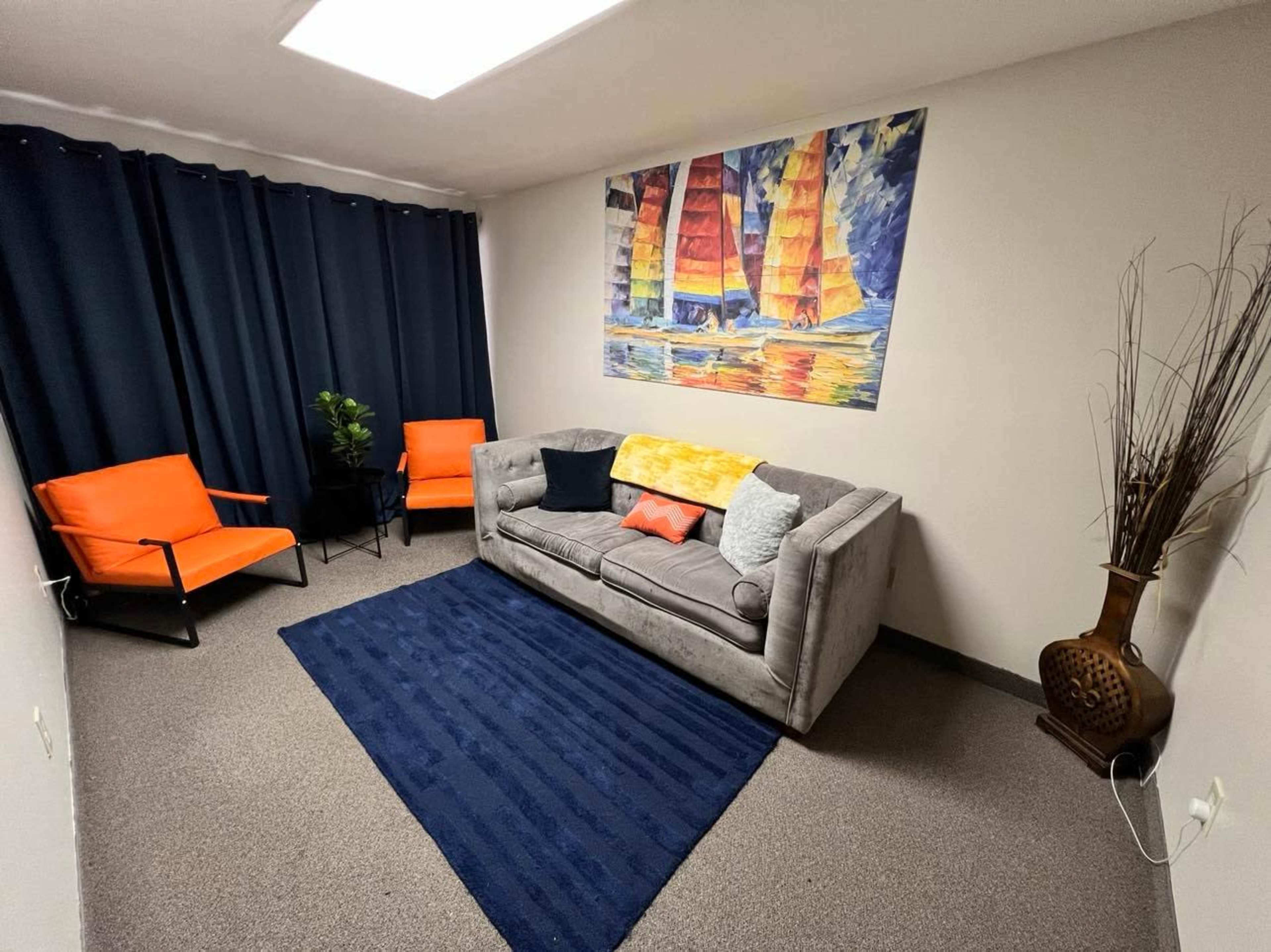 A small room furnished with a gray sofa, two orange chairs, a dark blue rug, and a colorful painting of sailboats on the wall, surrounded by black curtains.