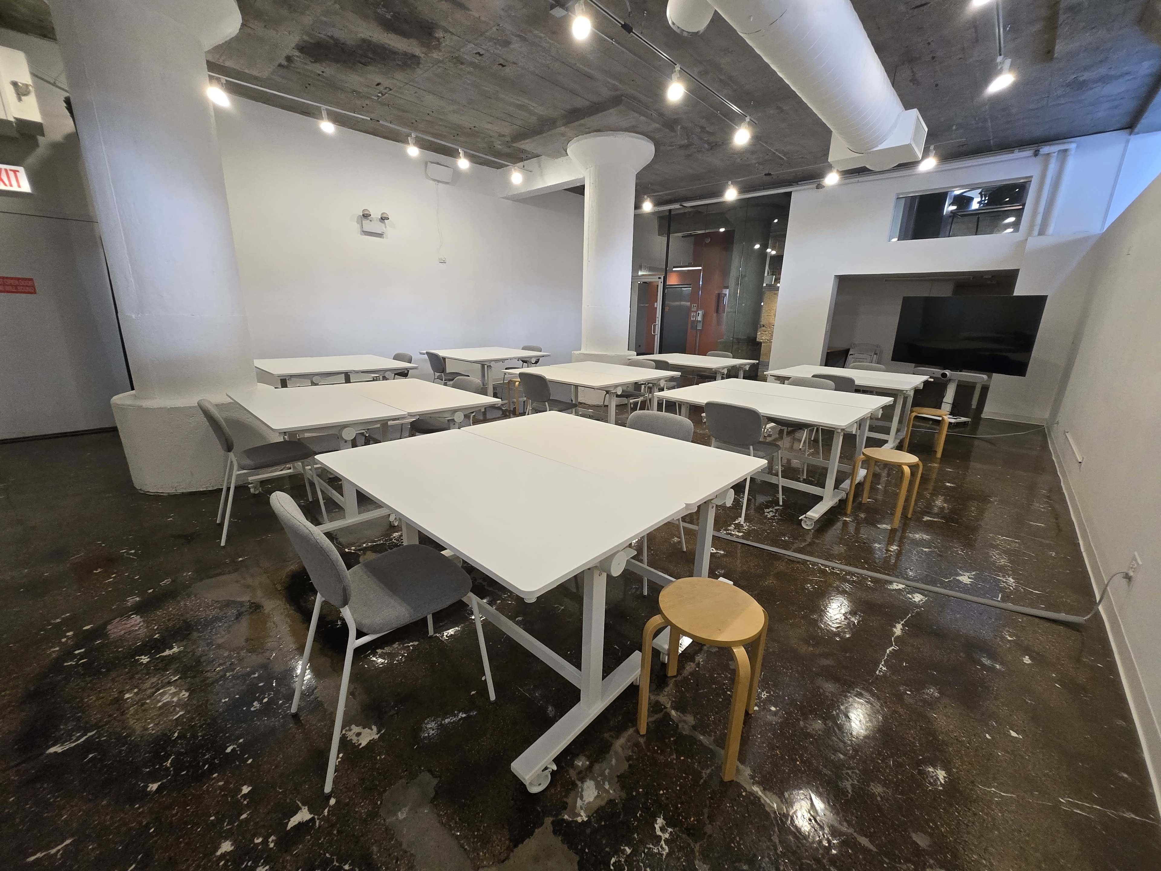 The image shows a large, empty meeting room with several white tables and chairs, and polished concrete floors beneath overhead lights.