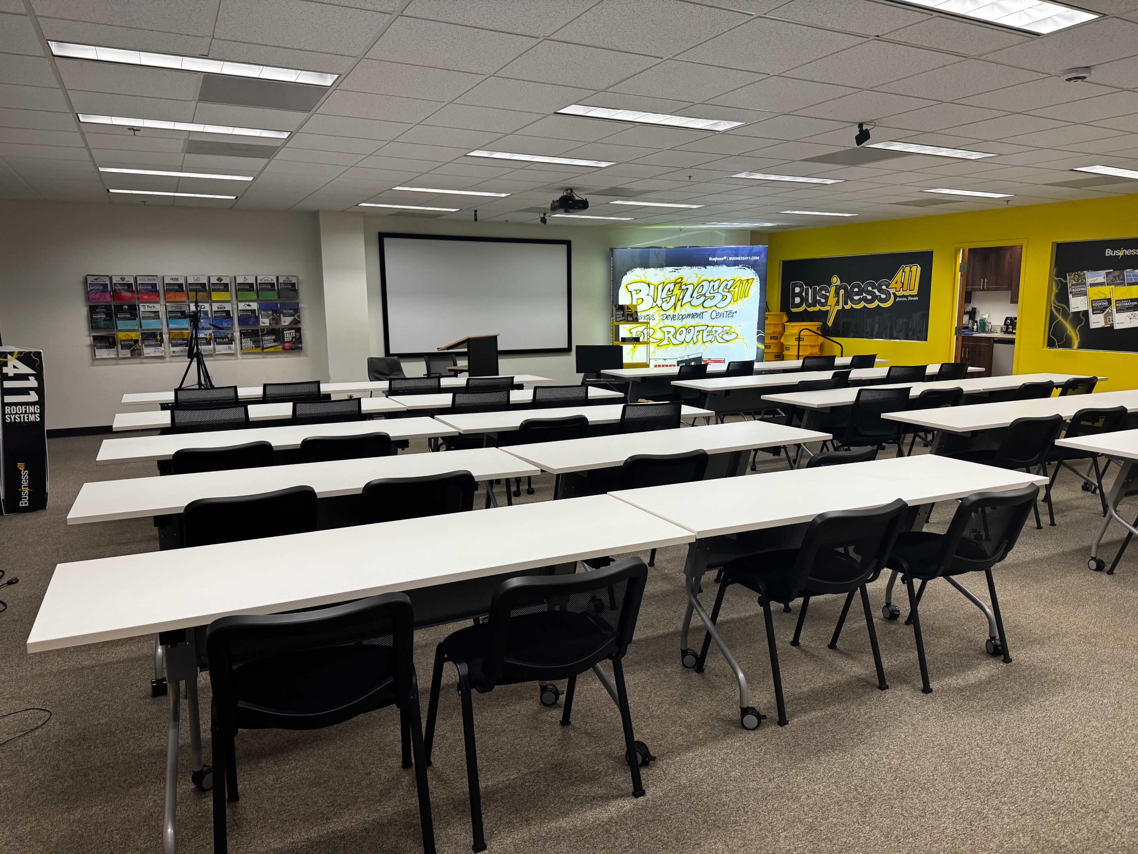 The image shows a classroom or training center with multiple rows of tables and chairs set up, a presentation screen at the front, and a projector hanging from the ceiling.