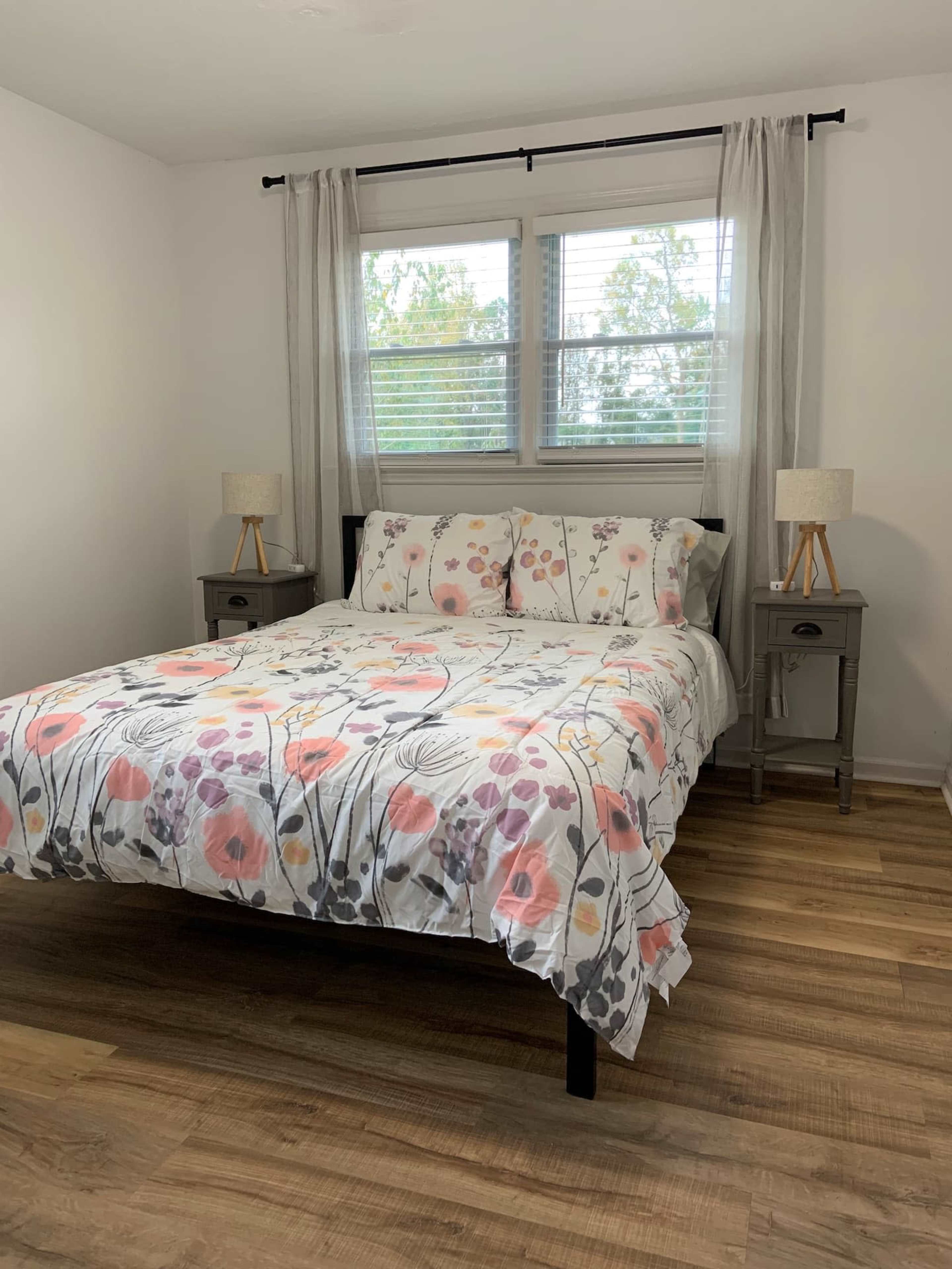 A minimalist bedroom featuring a bed with a floral-patterned duvet, two nightstands with lamps, and large windows allowing natural light to enter the room.