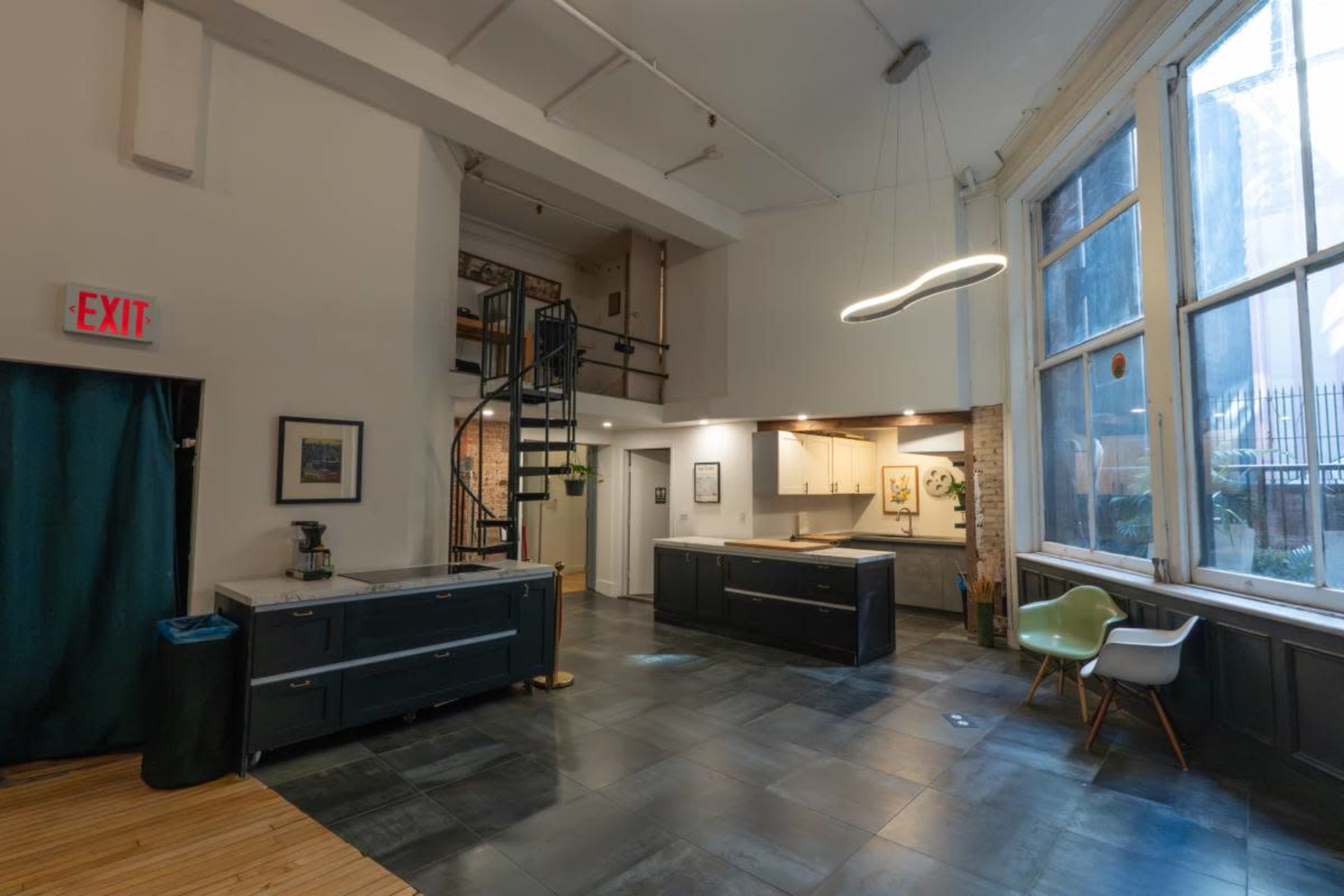 The image shows a spacious, modern interior featuring a kitchen area, large windows, a spiral staircase, and a mix of dark cabinetry and light flooring.