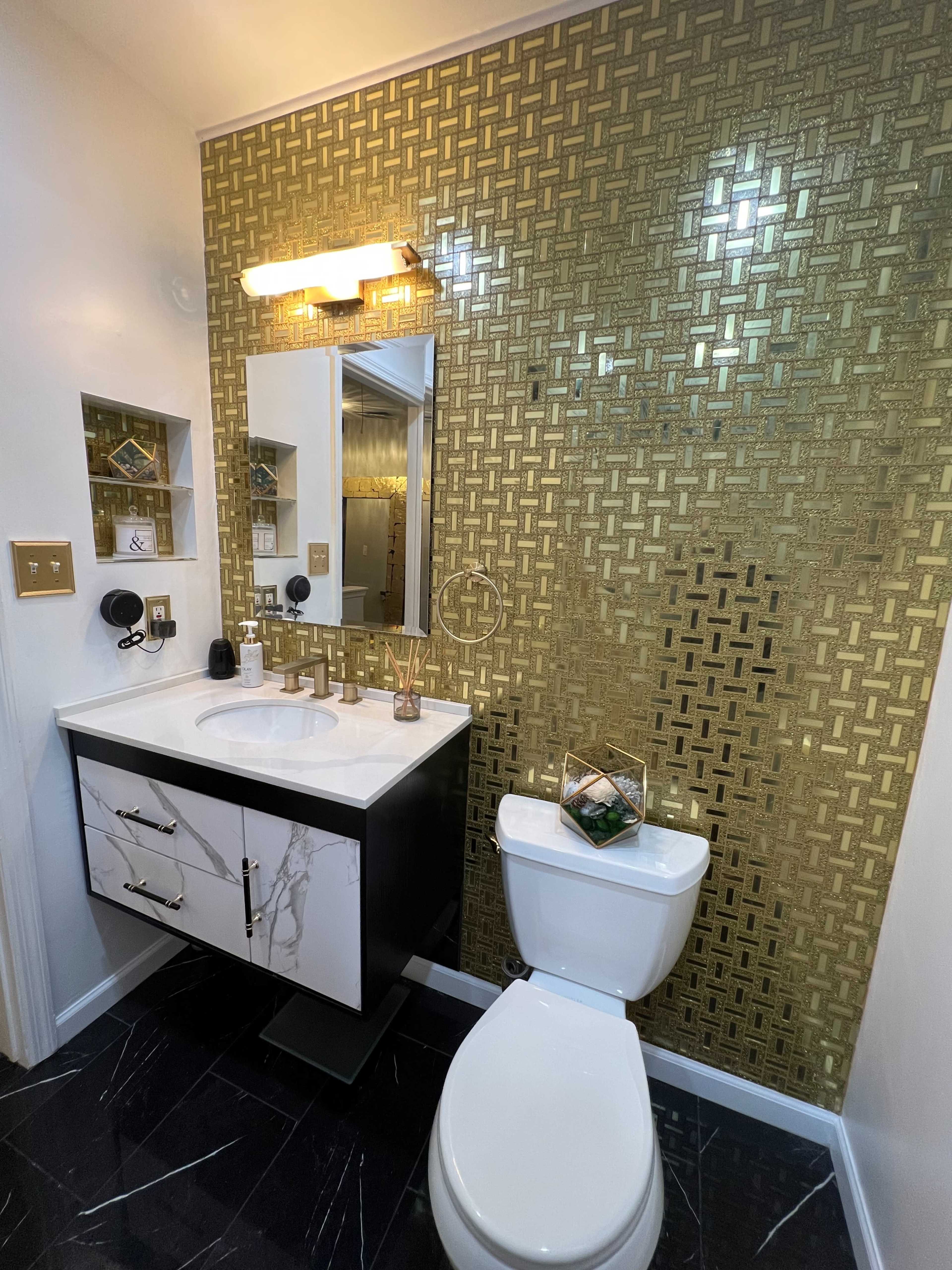 A modern bathroom featuring a gold patterned wall, a white sink with a black cabinet, a mirror, and a toilet.