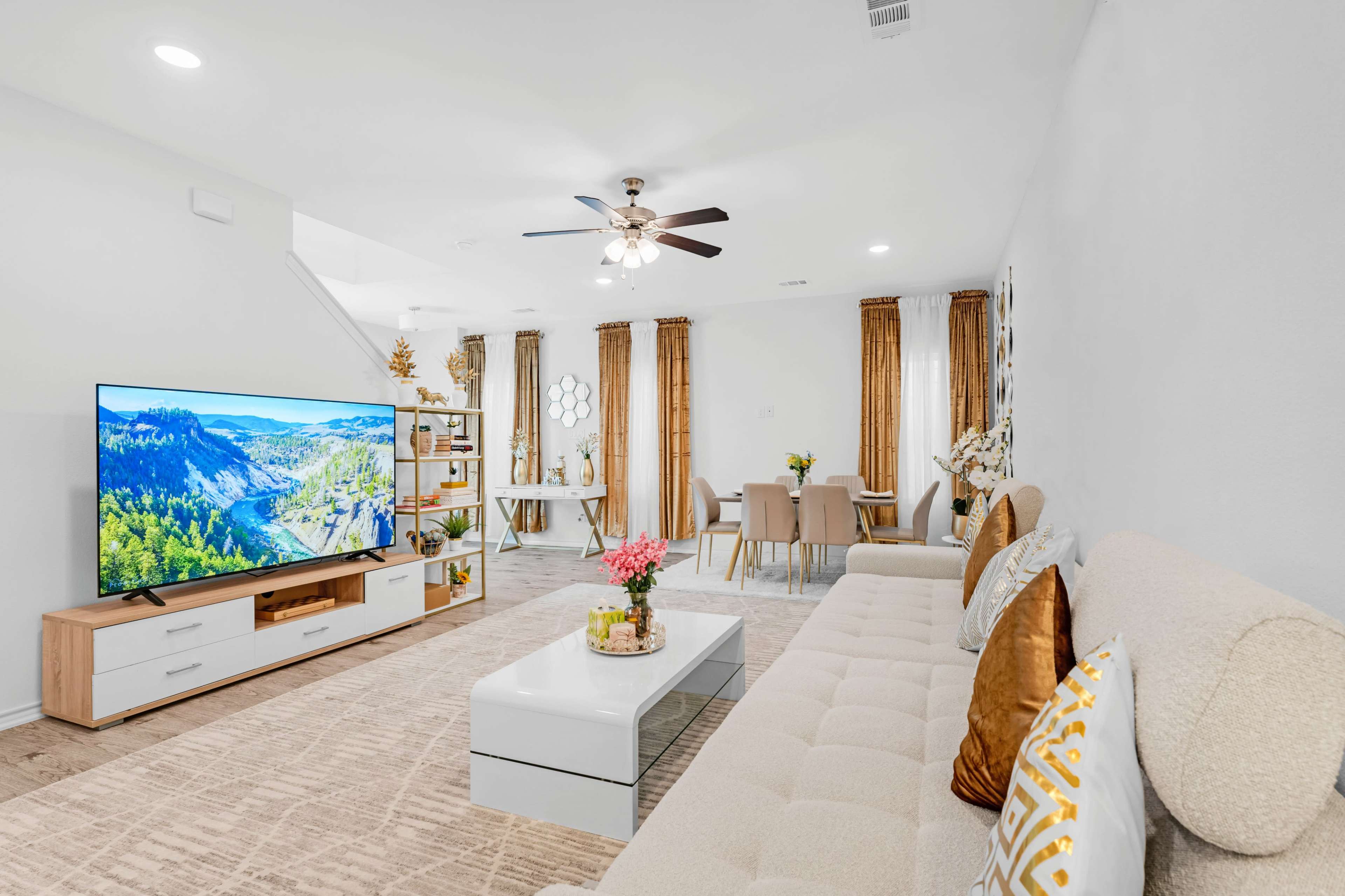 A modern living room features a large TV, a beige sectional sofa, a coffee table, and a dining area with a light-colored table set.