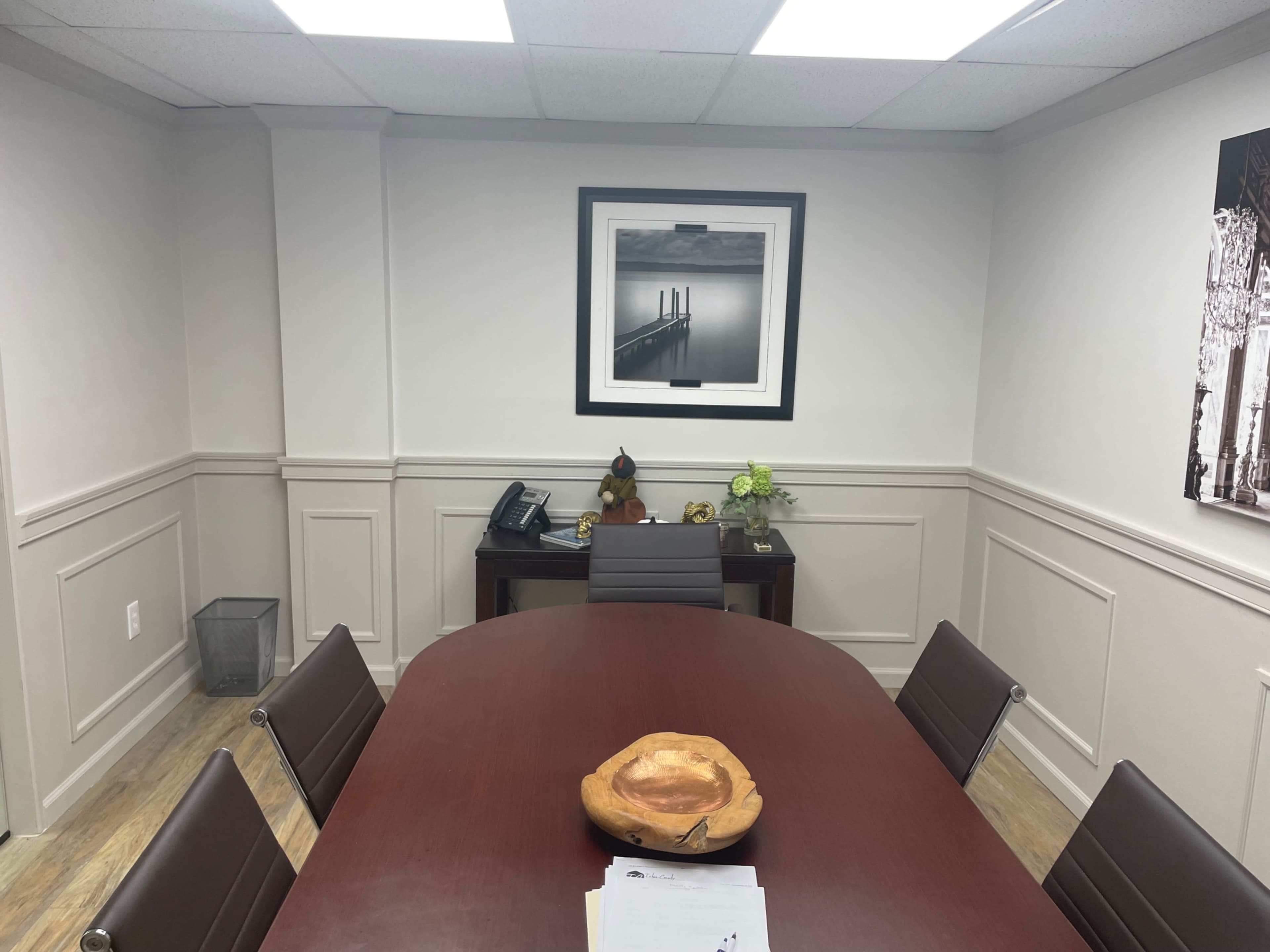 A conference room features a large oval table surrounded by six chairs, with a framed black-and-white photograph on the wall and a decorative bowl in the center of the table.