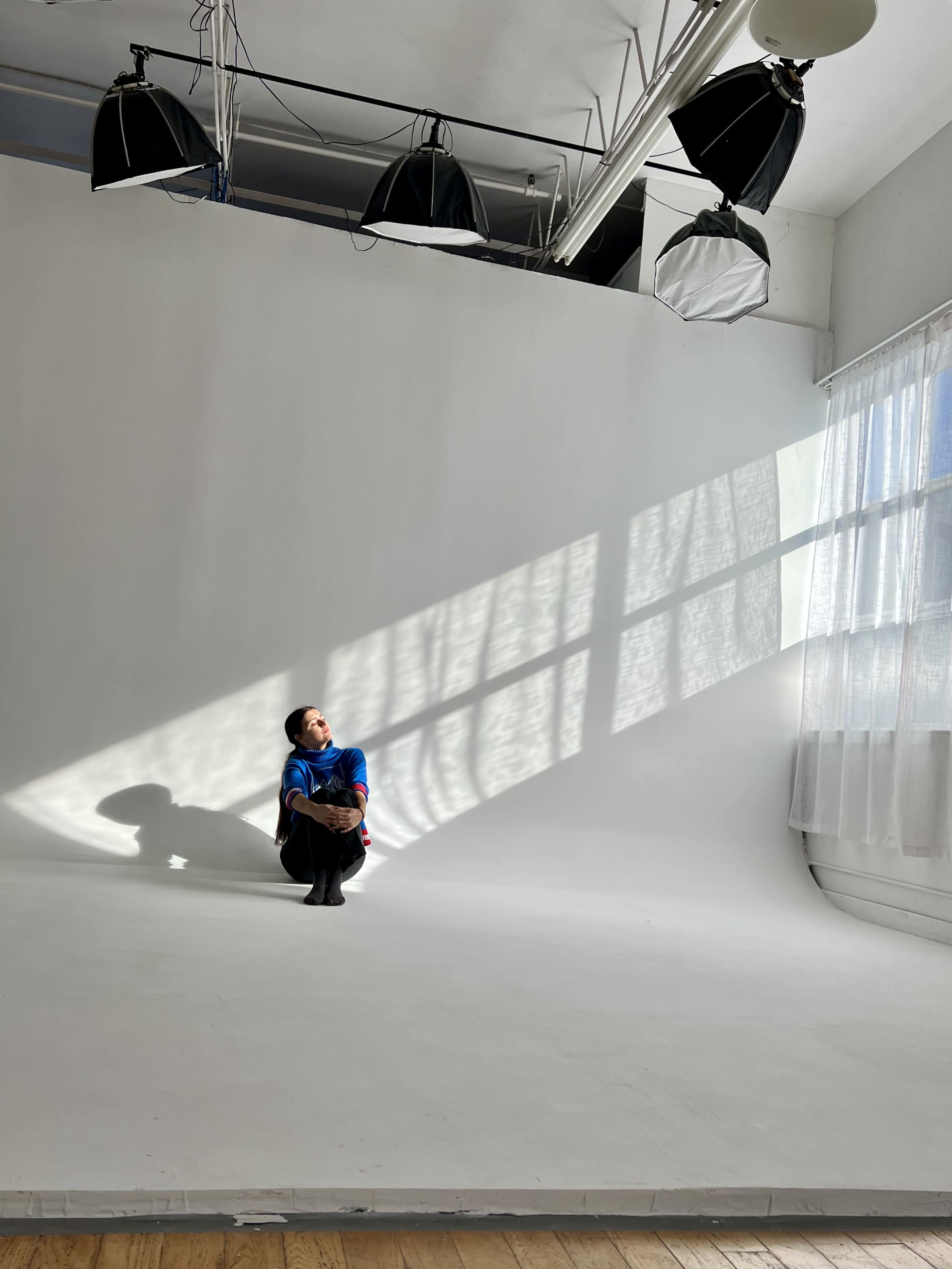 A person sits on the floor of a minimalist studio, illuminated by natural light streaming through a large window, casting geometric shadows on the white background.