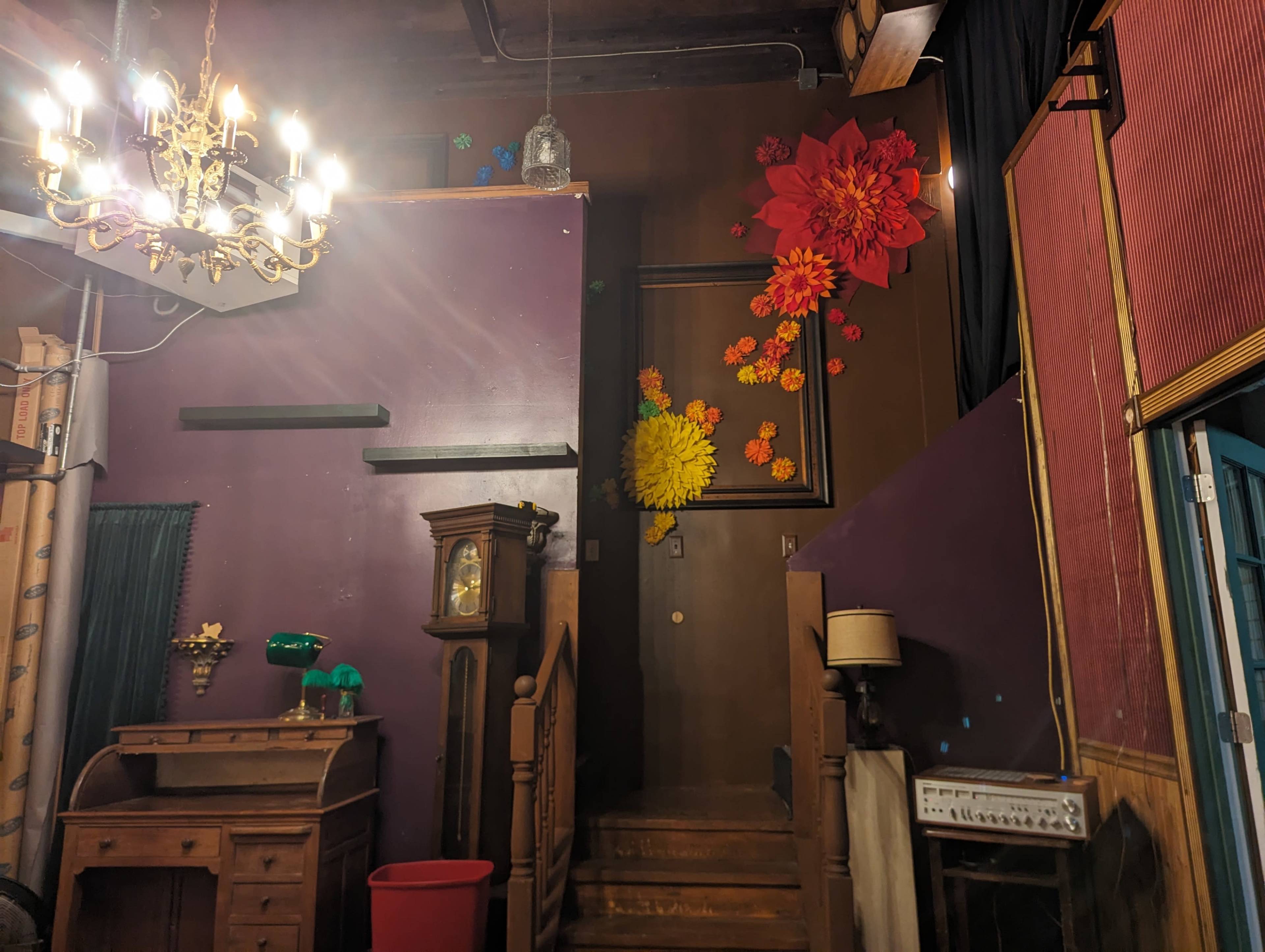 A dimly lit room with a staircase leading to a door, decorated with large paper flowers on the wall and a chandelier above.
