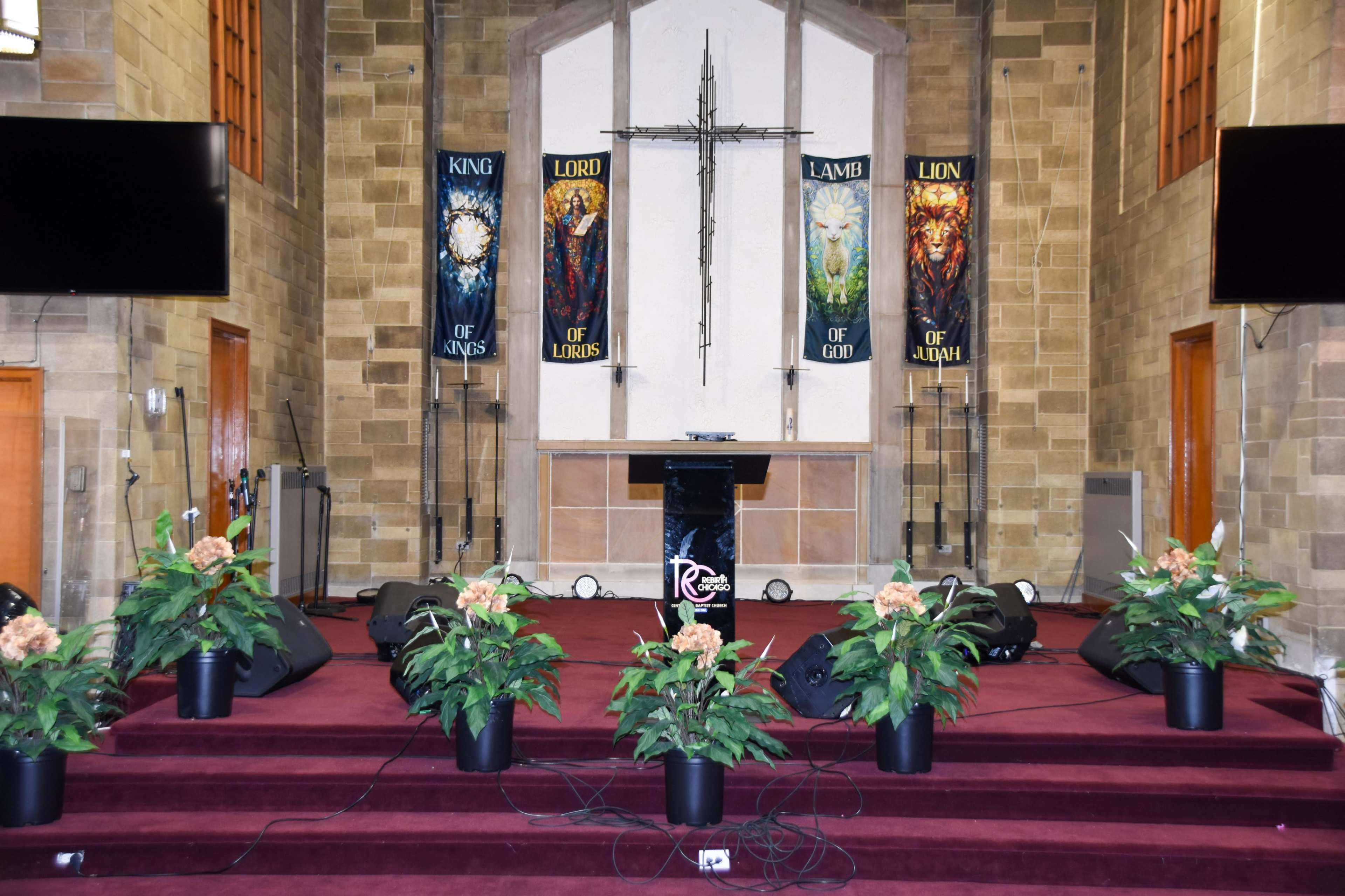 The image shows a church altar area with a cross, banners depicting biblical themes, a podium, and potted plants arranged on a red carpeted stage.