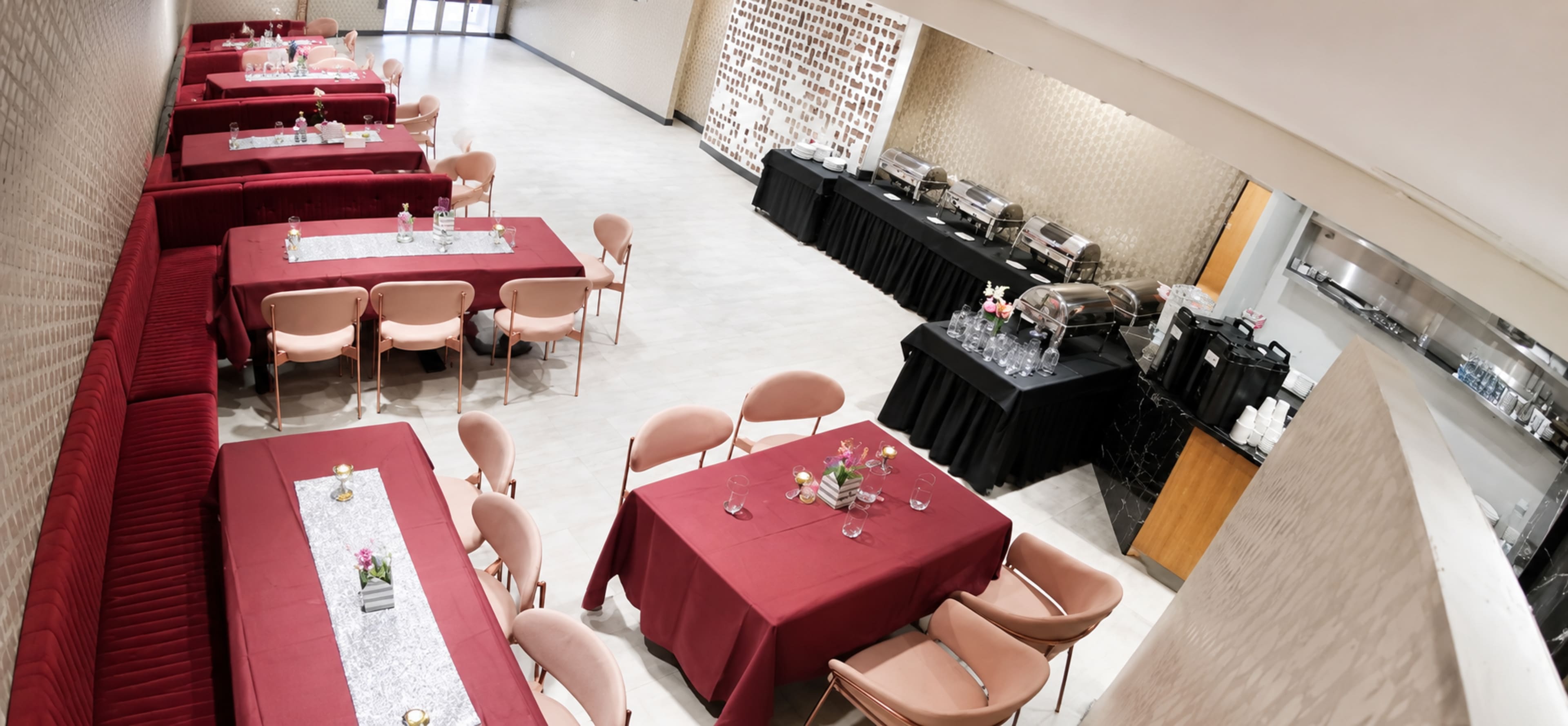 The image shows a spacious restaurant setting with neatly arranged tables covered in red tablecloths, featuring a buffet area and decorative floral centerpieces.