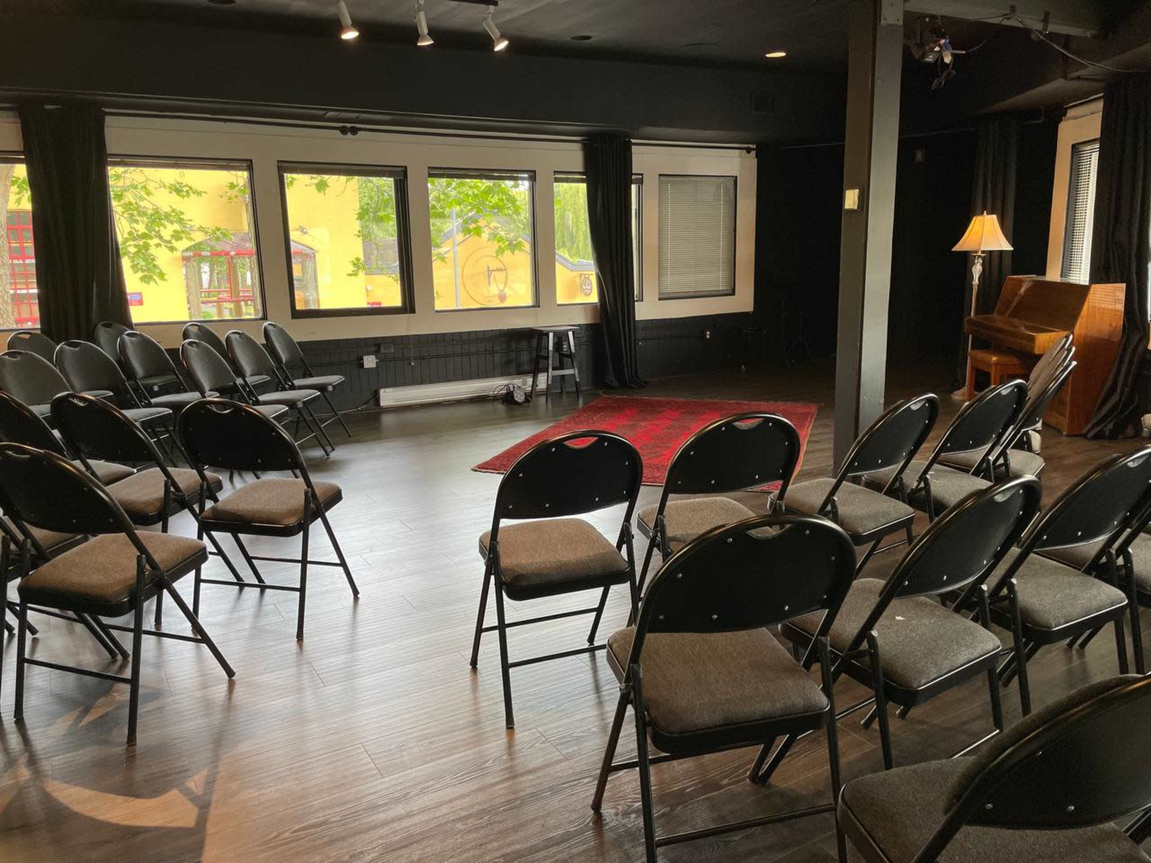 A room with black walls features a dozen black folding chairs arranged in rows facing a small stage area with a red rug and a piano.