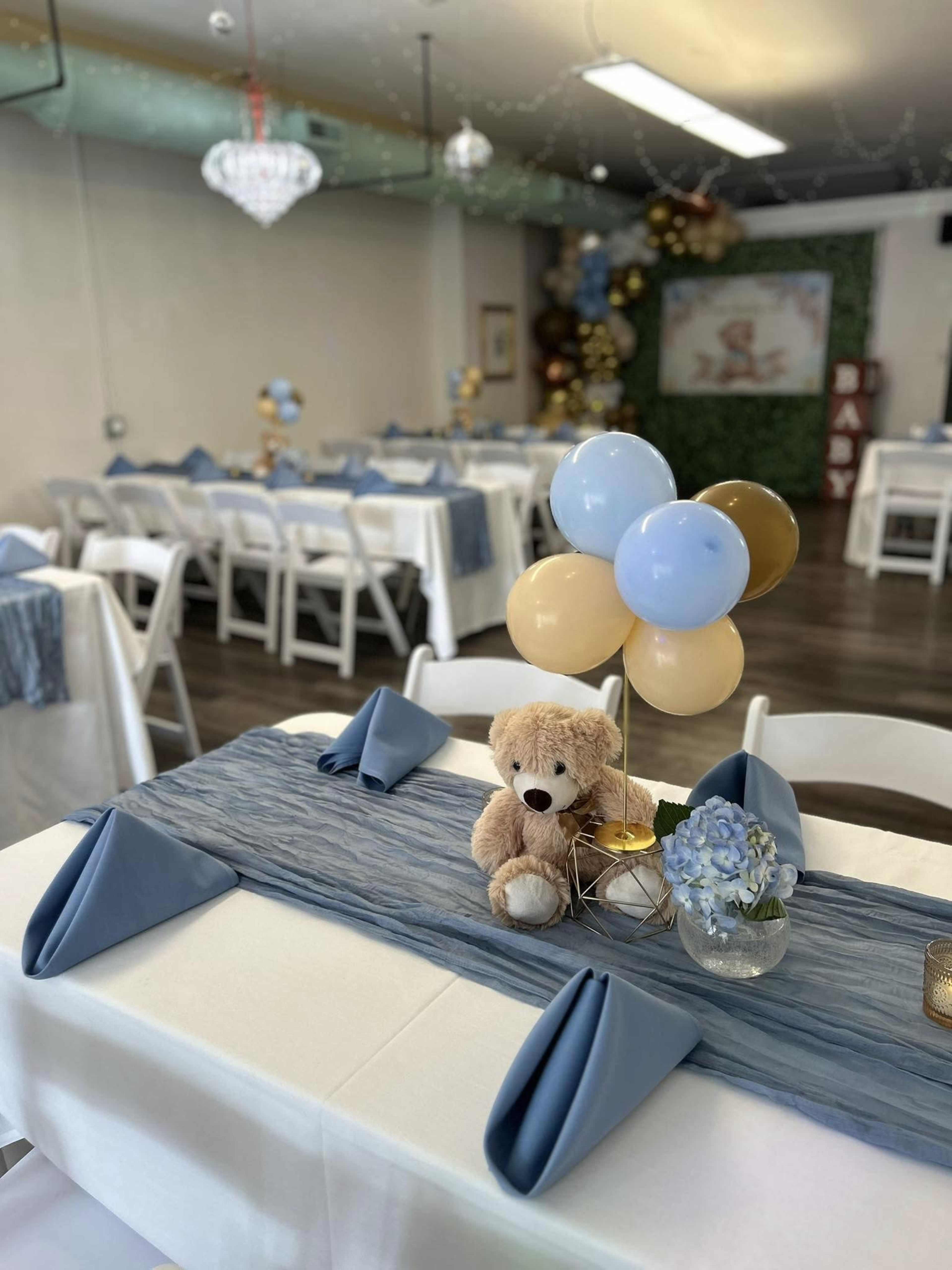 A table is set with blue and gold decorations, featuring a plush teddy bear and a bouquet of balloons, in a room prepared for an event.
