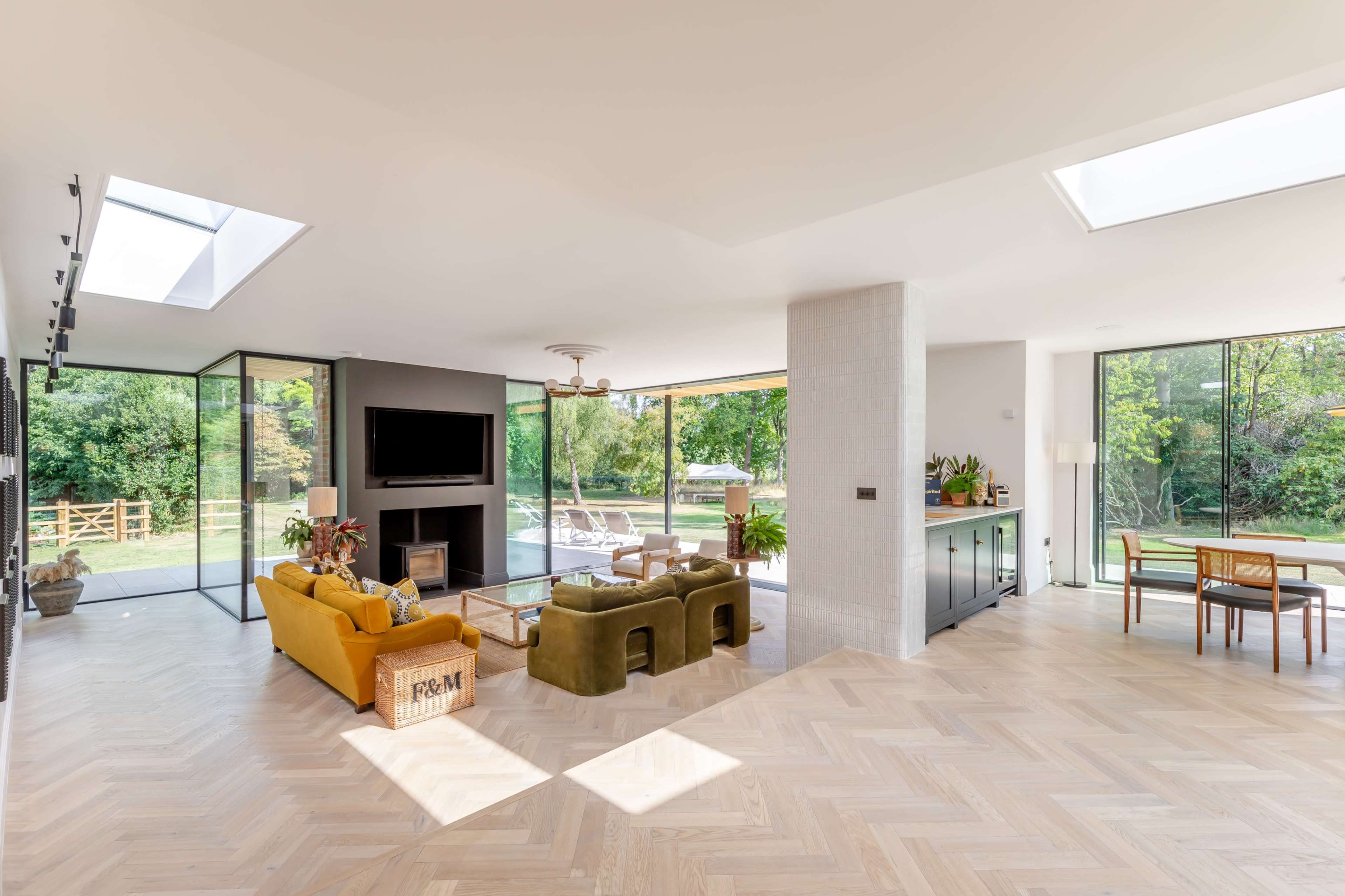 The image shows a modern open-concept living space with large windows, a fireplace, and stylish furniture overlooking a garden.