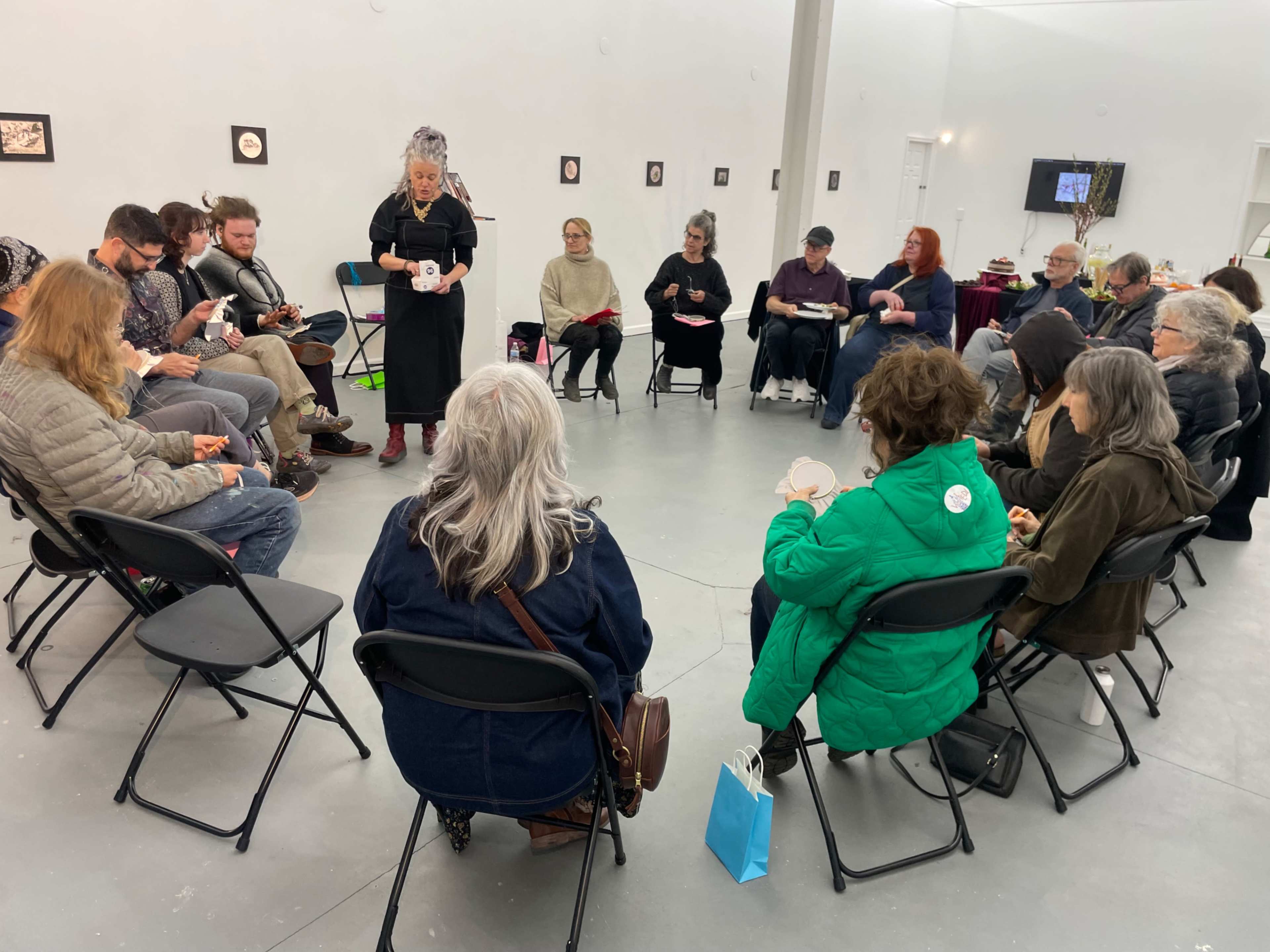 A group of people sits in a circle in an art gallery, attentively listening to a speaker in front of them.