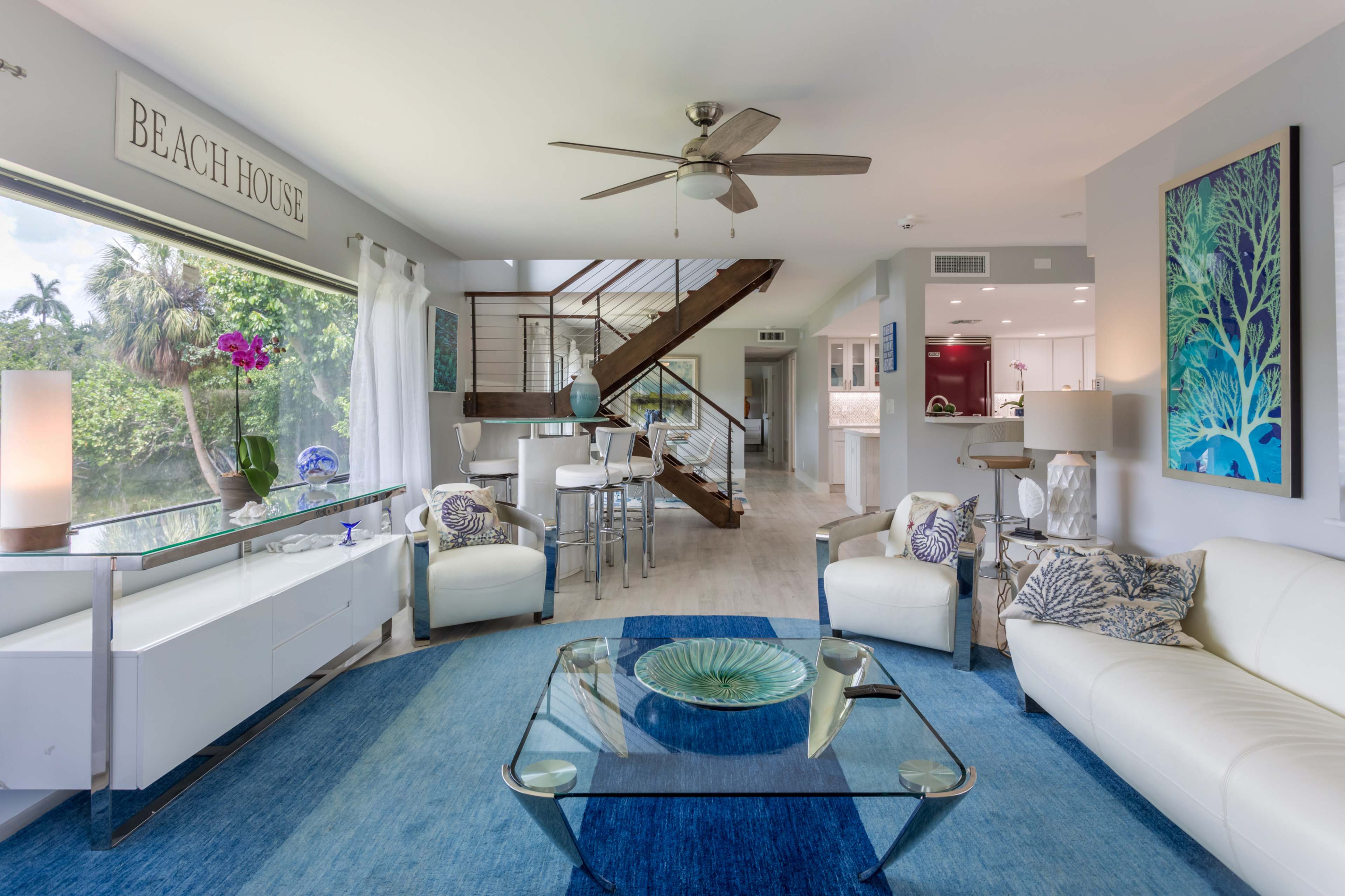 A bright living room features white furniture, a glass coffee table, and a large window overlooking a lush outdoor area.