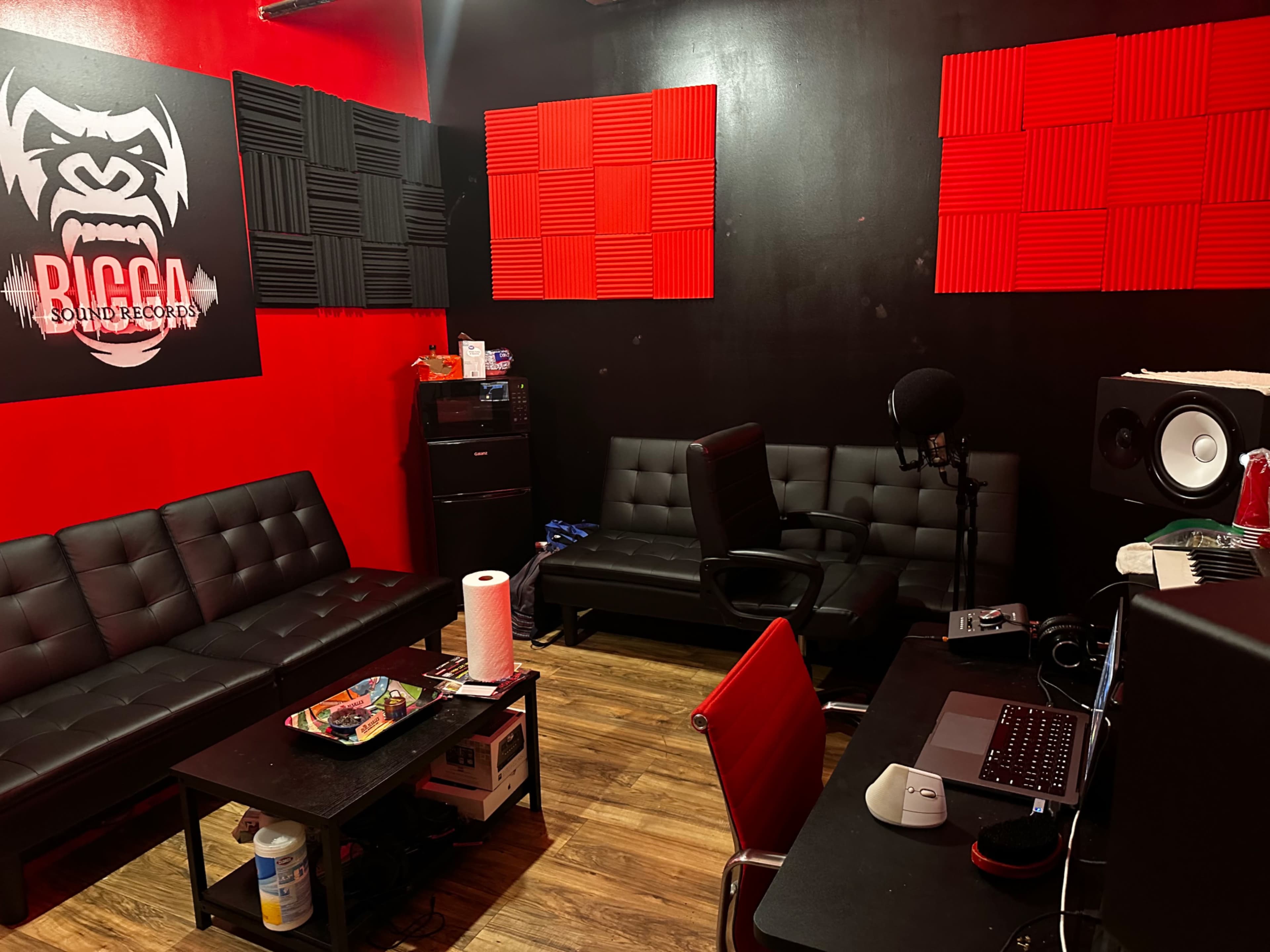 A music recording studio with black and red walls, featuring black couches, soundproof panels, a microphone, speakers, and a computer set up on a desk.