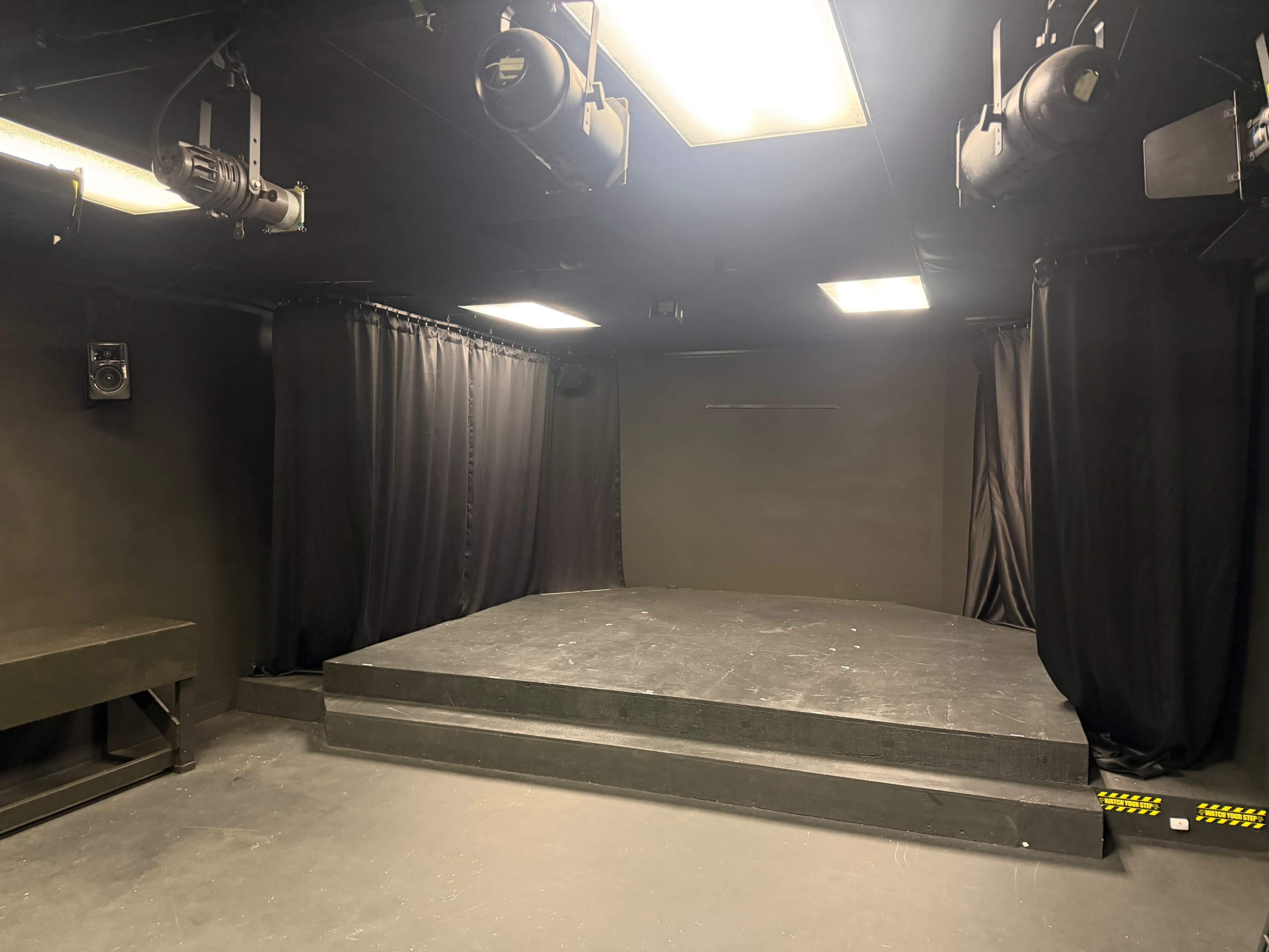 The image shows a small black box theater with a raised stage, black curtains, and overhead lighting fixtures.