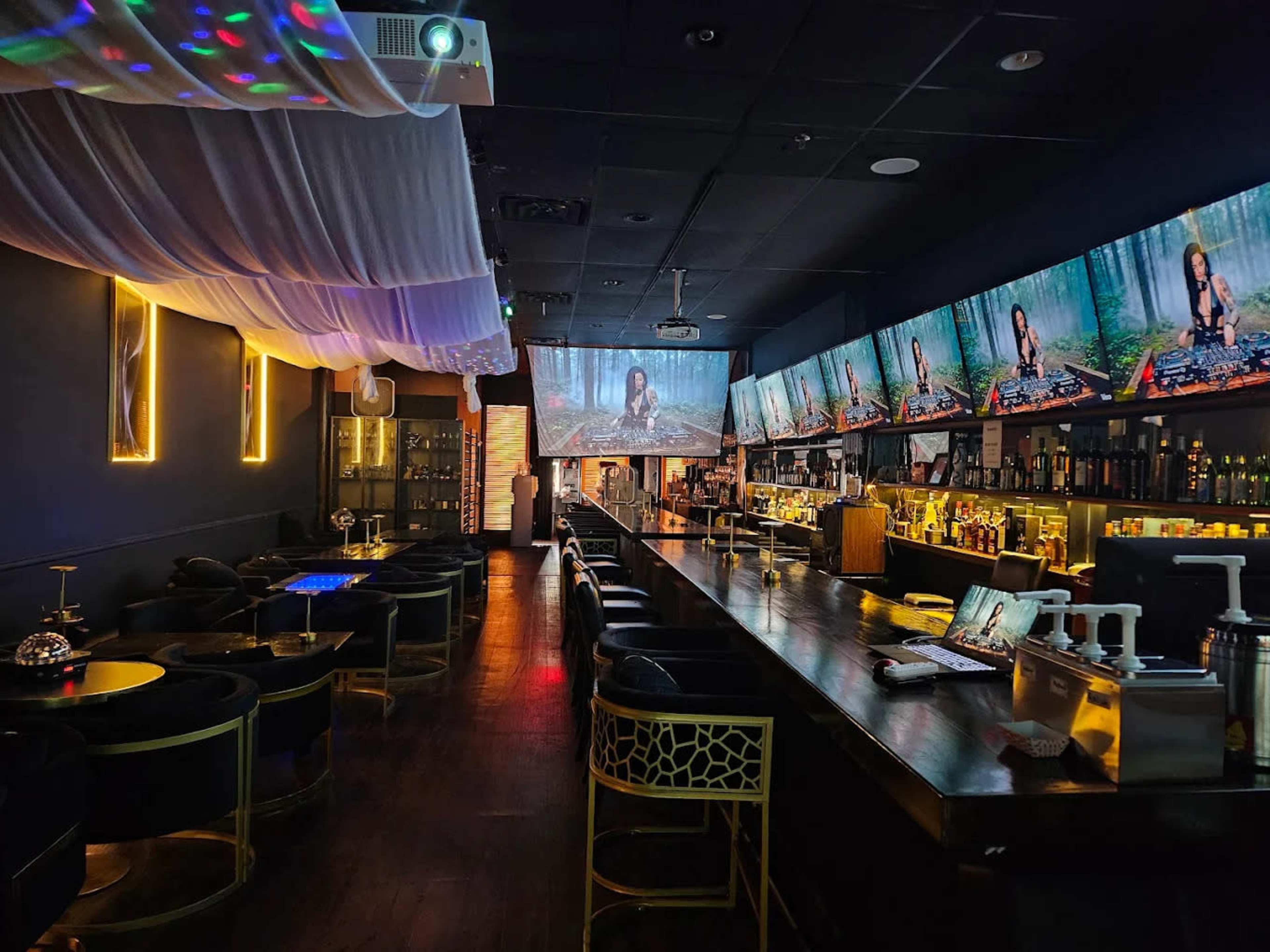 The interior of a dimly lit lounge features a long bar with multiple screens displaying visuals, surrounded by stylish seating and atmospheric lighting.