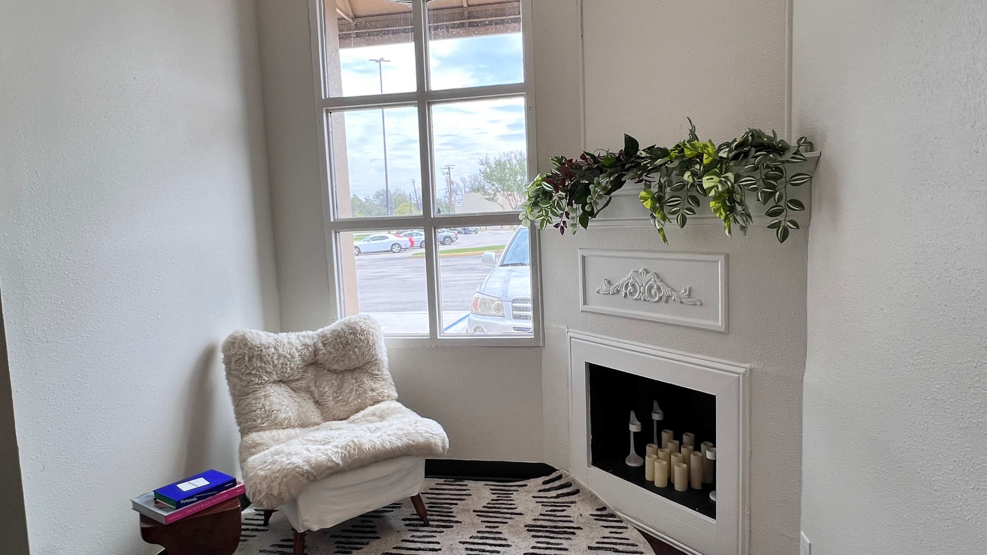A cozy corner features a plush chair beside a decorative fireplace with candles and a window adorned with greenery.