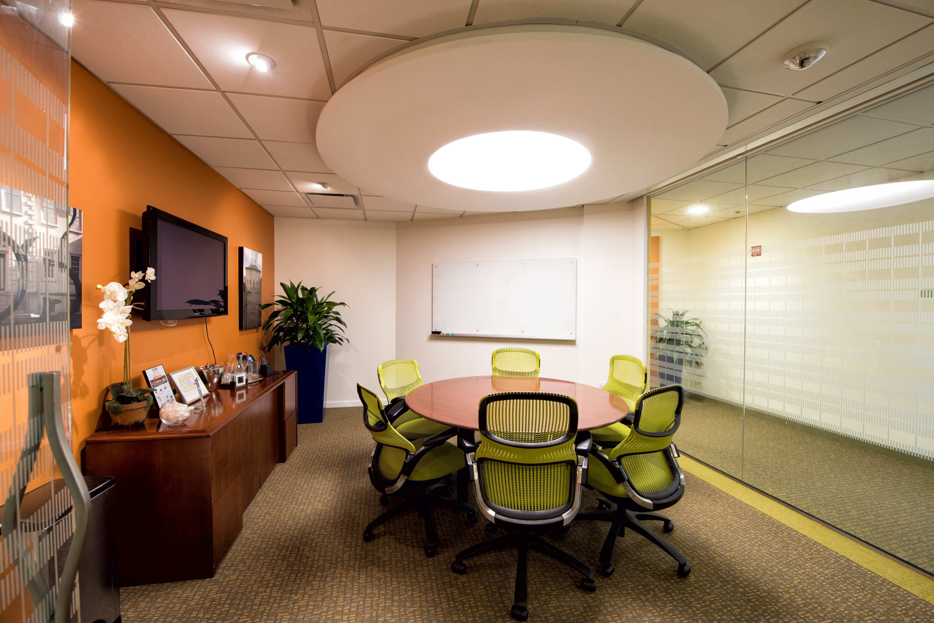 A modern conference room with a round wooden table surrounded by six green office chairs, a whiteboard, and a television on the wall.
