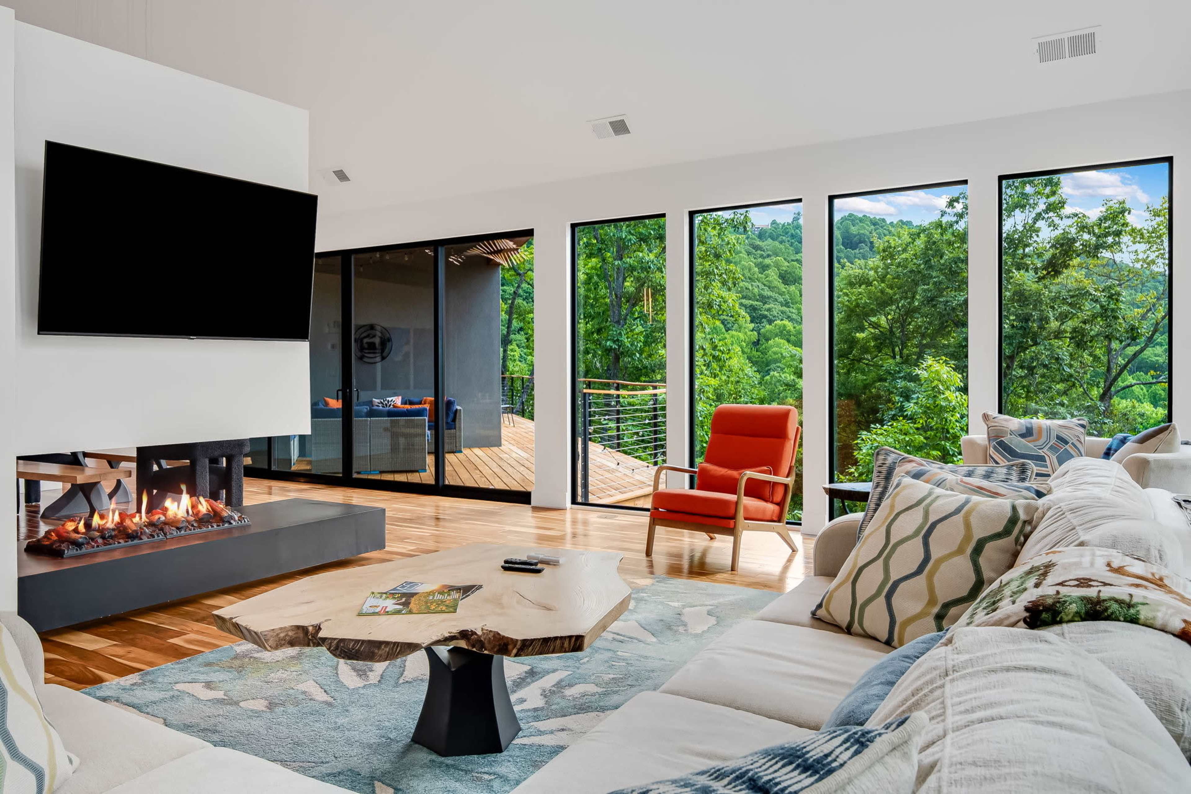 A modern living room features a fireplace, large windows overlooking greenery, and contemporary furnishings, including a red chair and a natural wood coffee table.