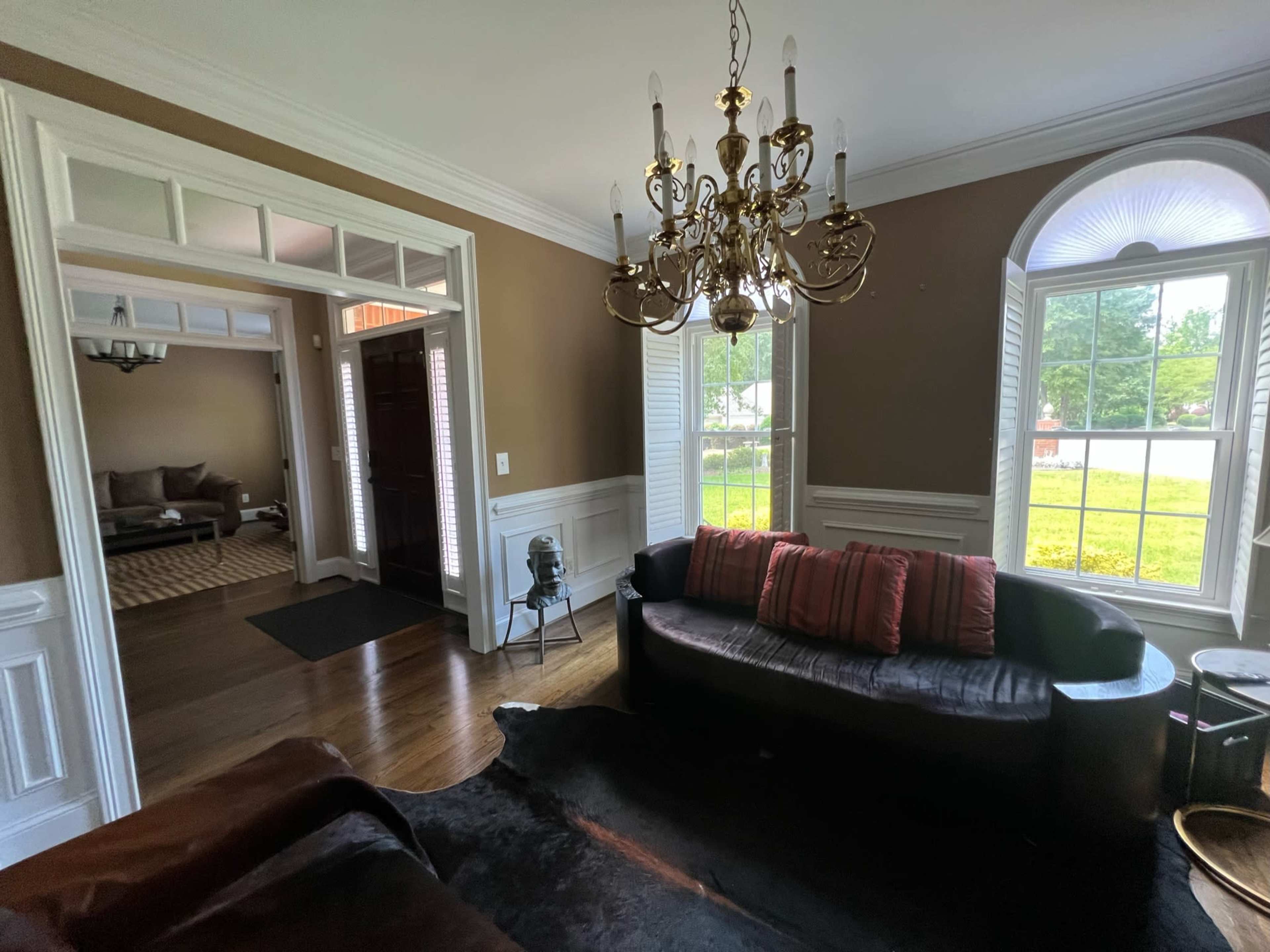 A living room with a chandelier, a black sofa adorned with cushions, and large windows letting in natural light.