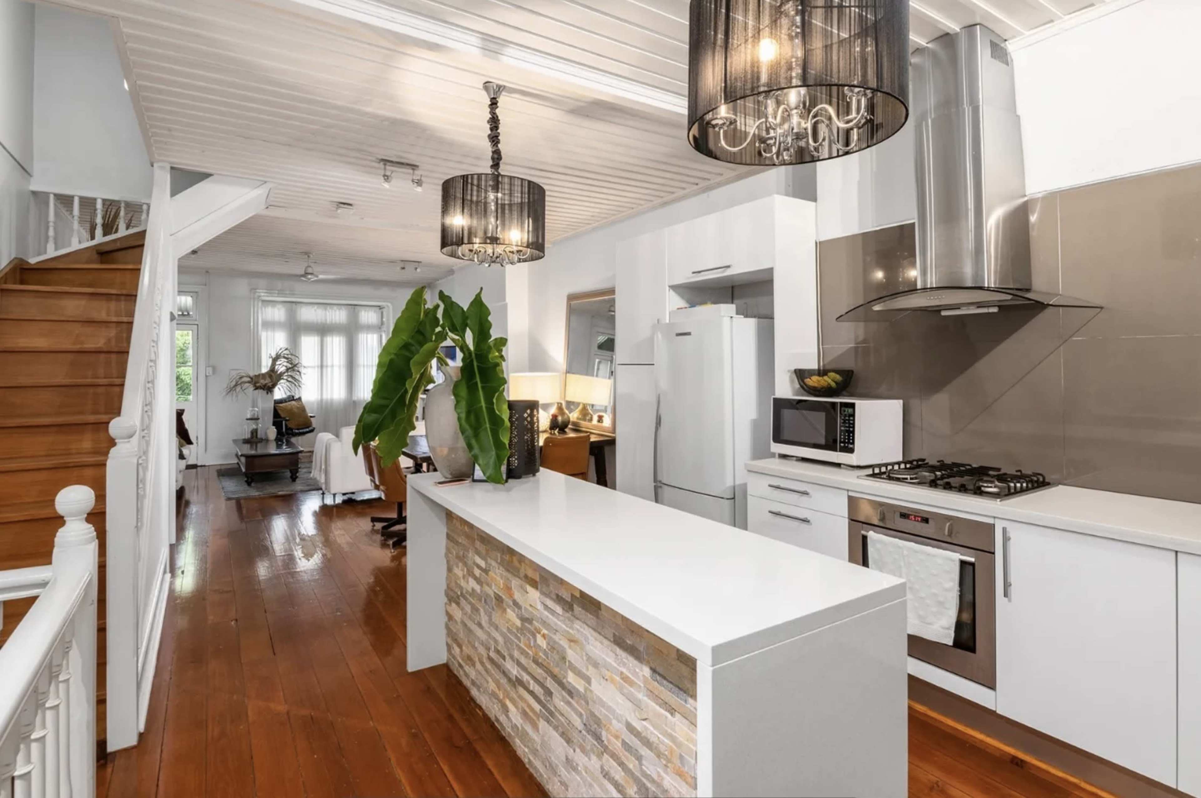 The image shows a modern kitchen featuring a central island with a stone facade, stainless steel appliances, and pendant lighting, alongside an open living area with wooden flooring and a staircase.