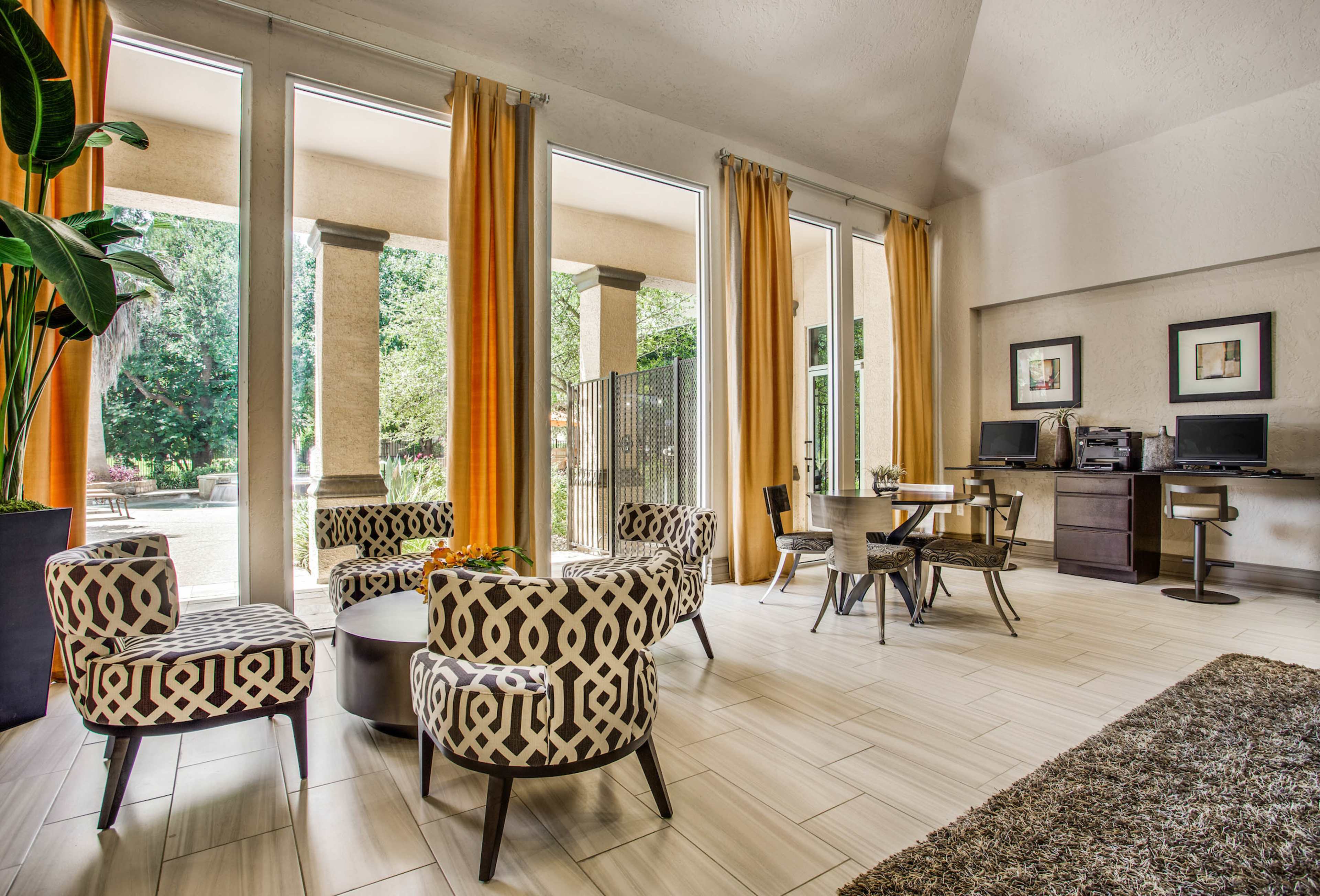 A spacious lounge area with patterned chairs, a round table, and workstations, surrounded by large windows and bright curtains.
