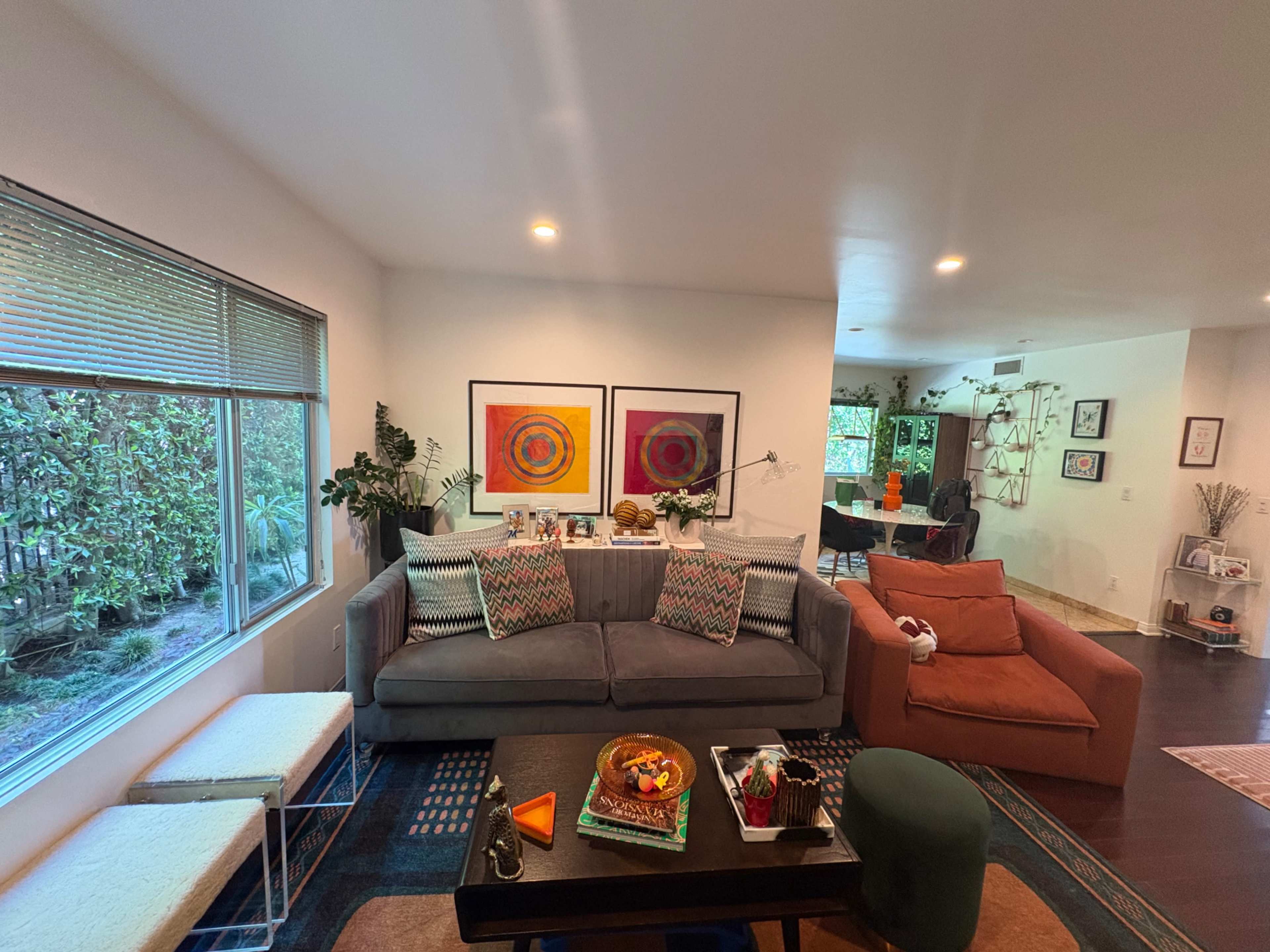 The image shows a brightly lit living room with a gray sofa, colorful art on the walls, and a view of greenery outside through a large window.