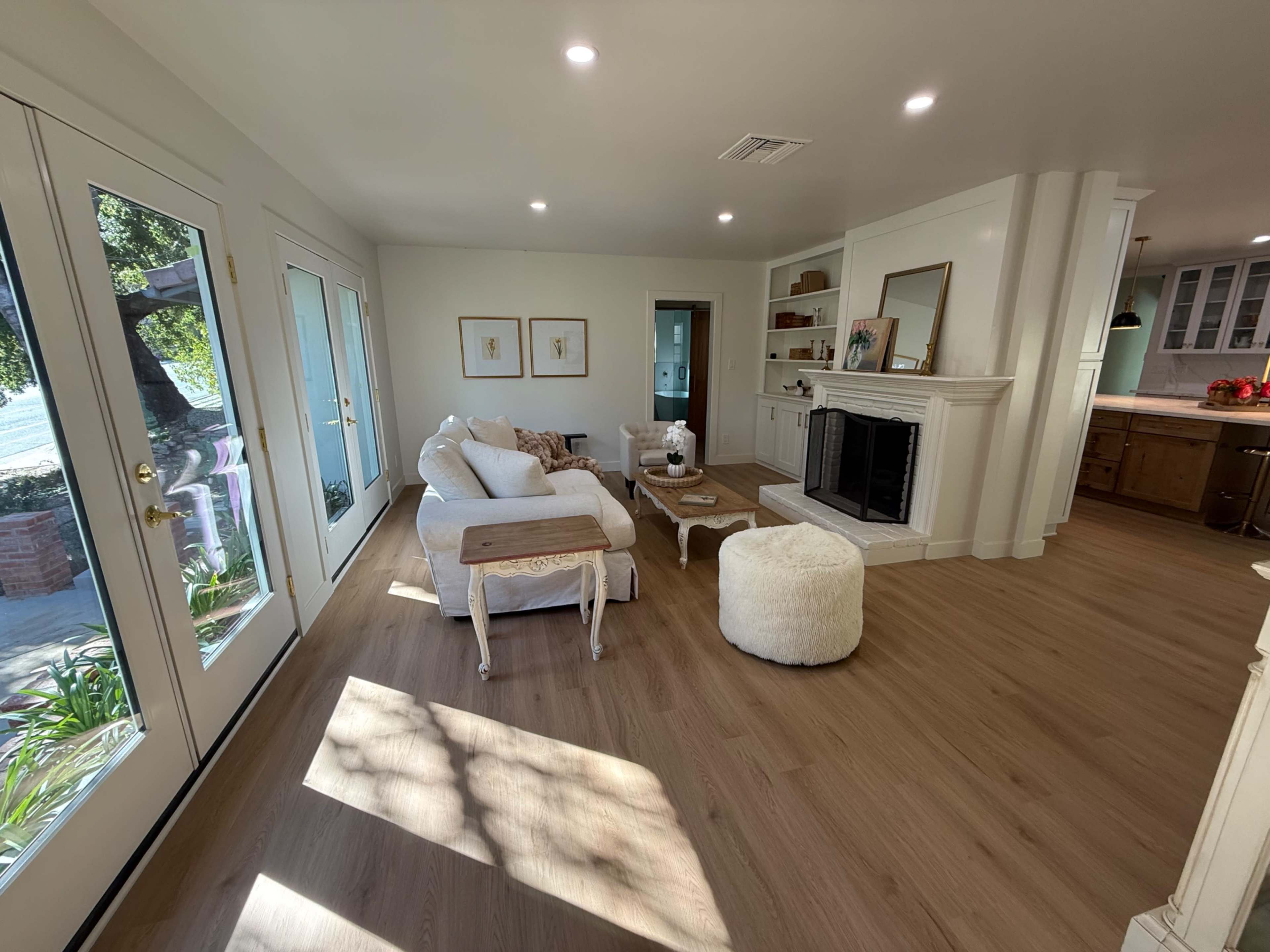The image shows a bright, modern living room featuring a white sofa, a round coffee table, and light wood flooring, with large windows allowing natural light to enter the space.