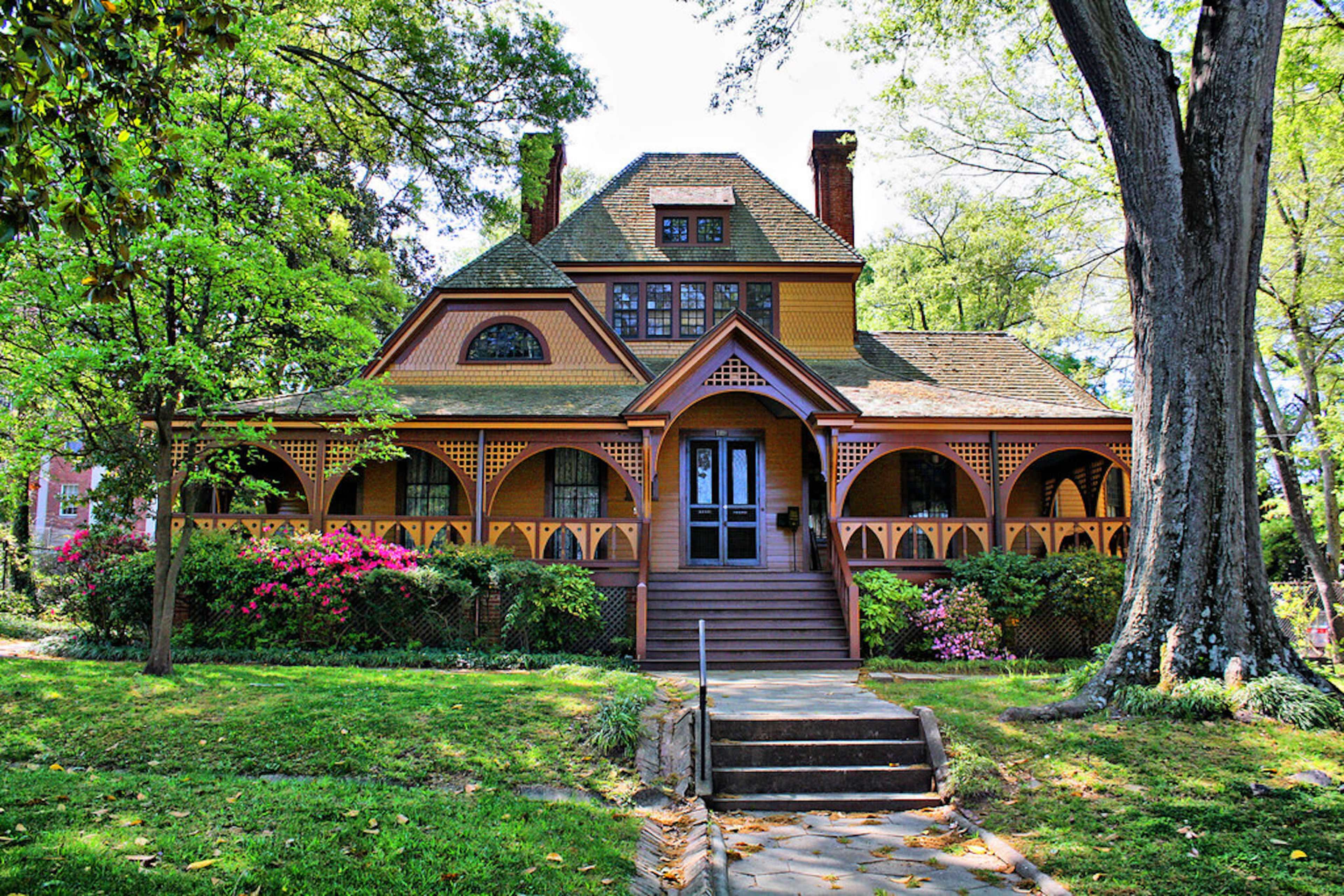 A large Victorian-style house with a wraparound porch and colorful flower beds sits surrounded by green trees and grass.