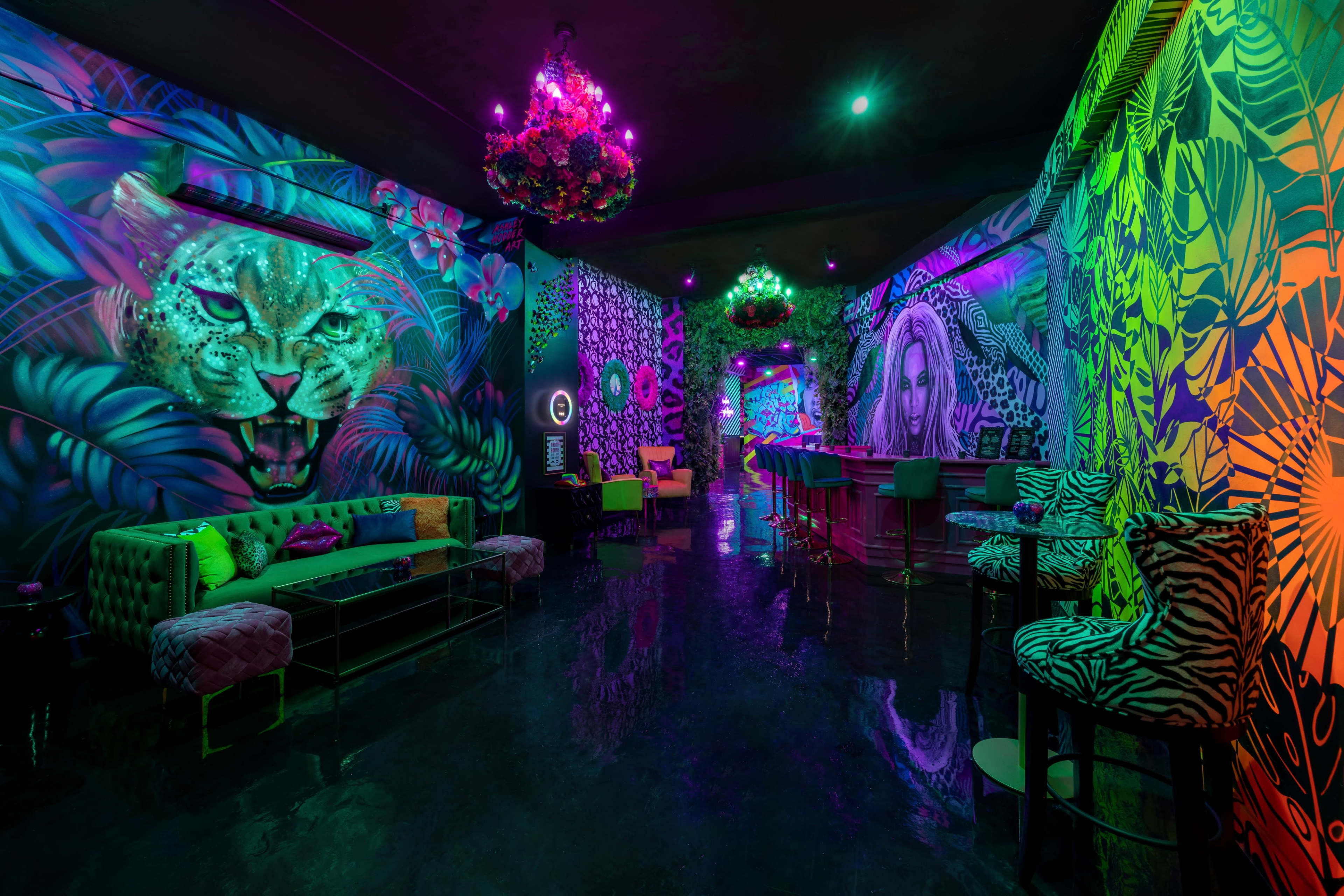 The image depicts a vibrant lounge with neon murals of a leopard and foliage on the walls, and brightly colored furnishings, all illuminated under purple and green lighting.