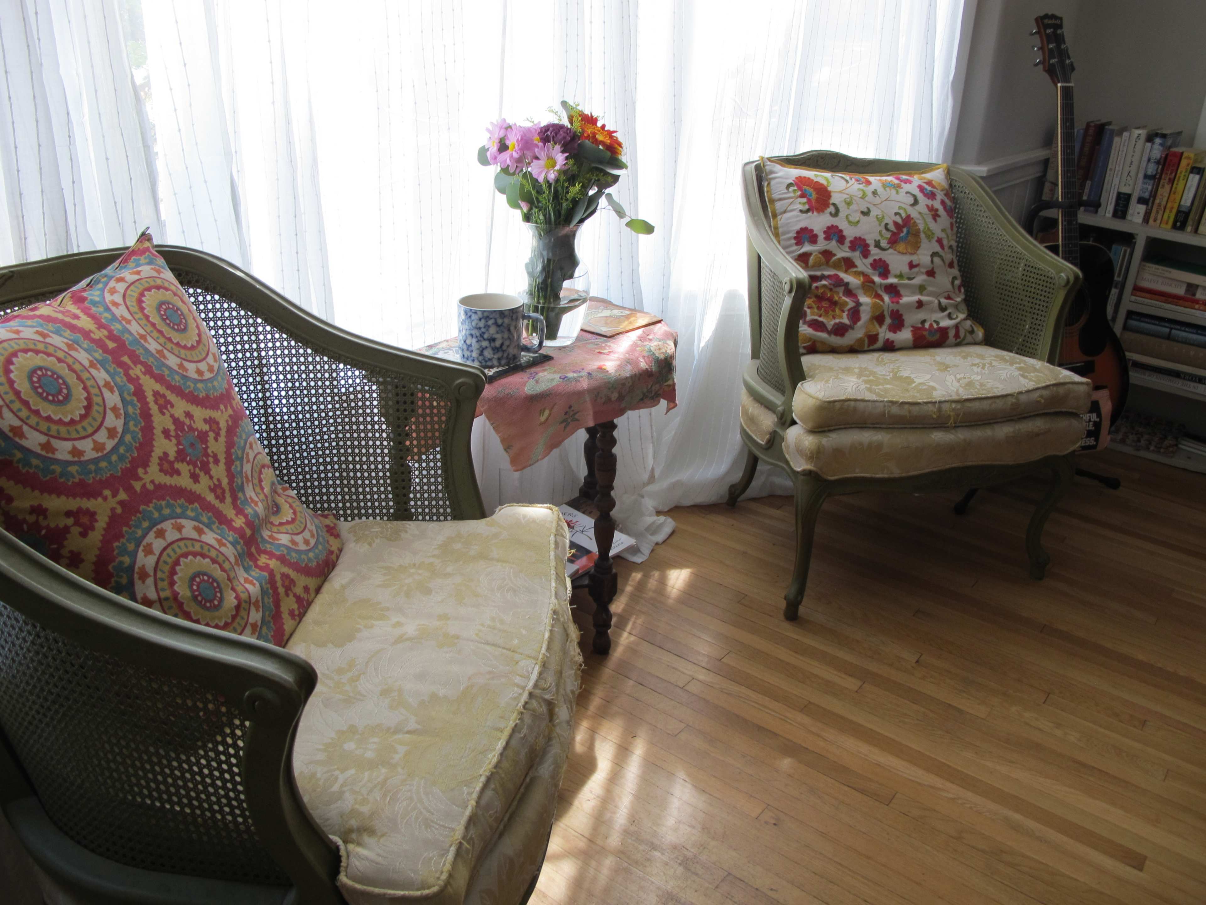 Two caned armchairs with colorful cushions are positioned next to a wooden side table adorned with a vase of flowers in a brightly lit room.