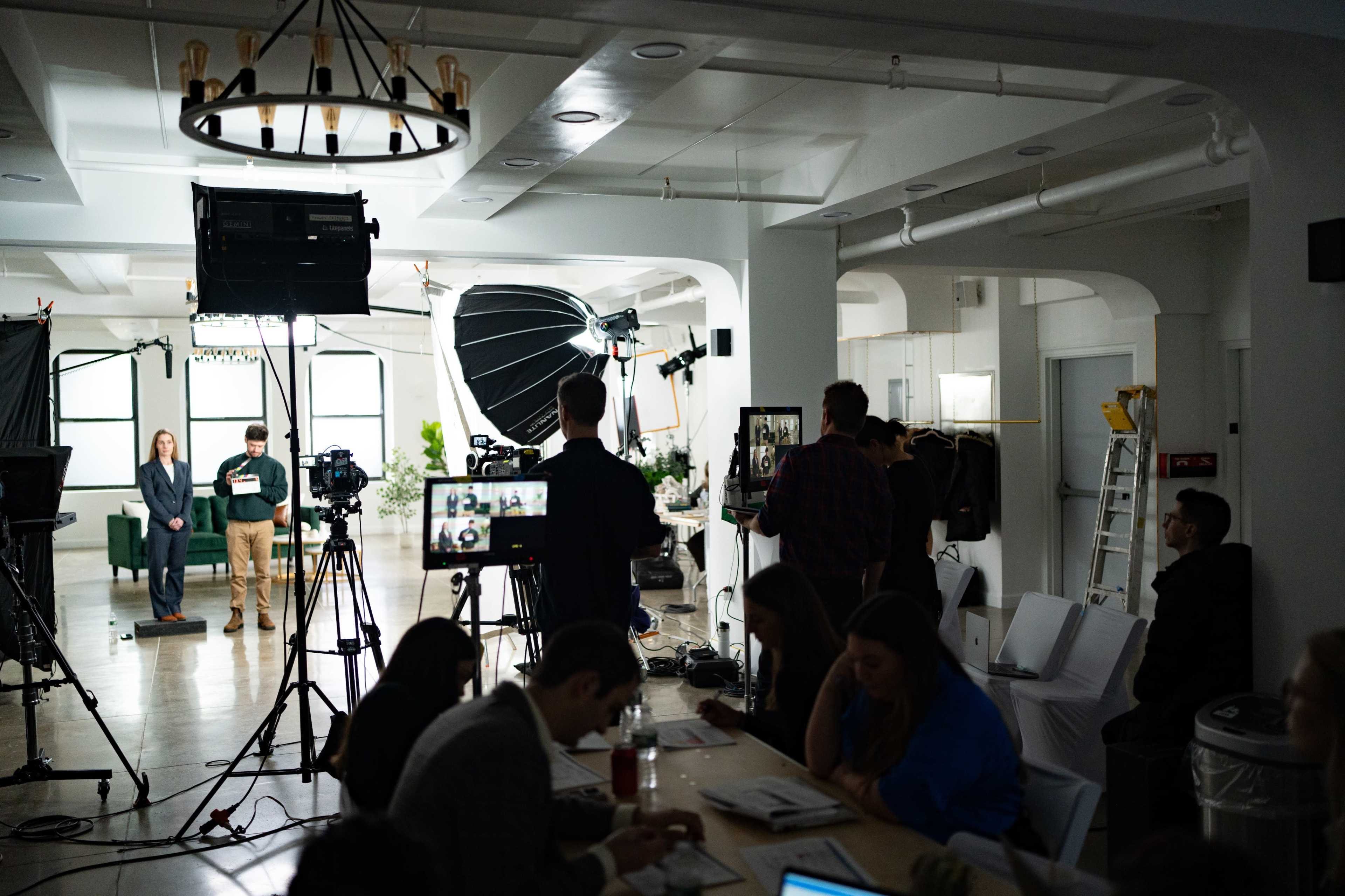 A film crew sets up for a recording session in a well-lit studio with various cameras and lighting equipment.